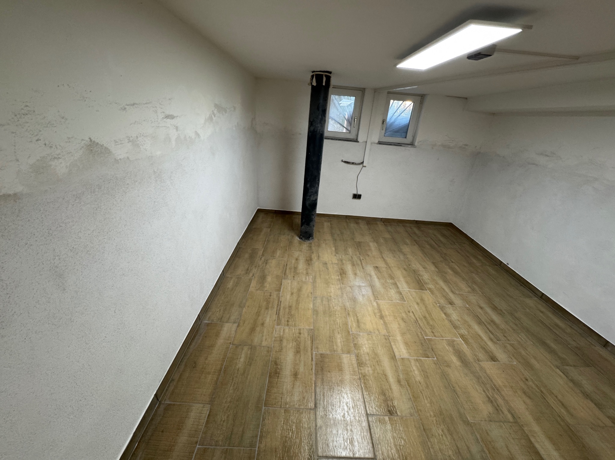 Wykończone wnętrze piwnicy z drewnopodobną podłogą, czarną rurą pionową i oknami w białych ramach, ściany częściowo przygotowane do malowania.