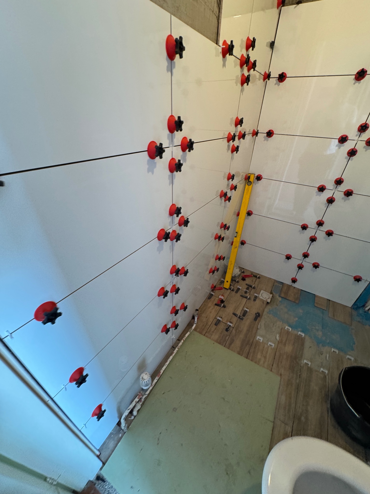 Układanie białych płytek w łazience z użyciem systemu poziomowania płytek (czerwone elementy) i poziomicy; widoczny częściowo zamontowany sedes i surowy stan podłogi.