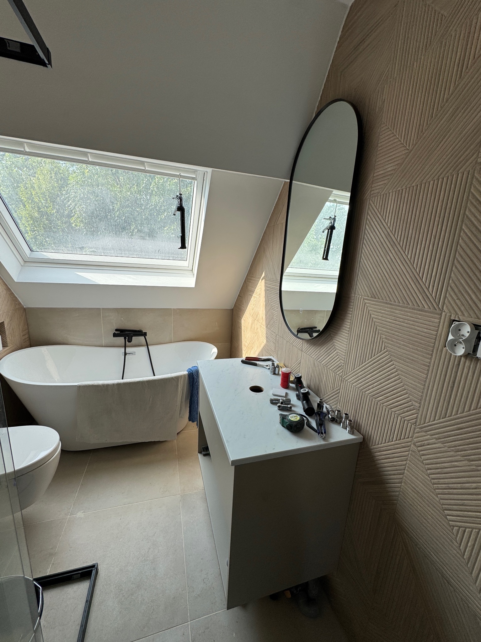 Nowoczesna łazienka na poddaszu z wanną wolnostojącą, umywalką z blatem i owalnym lustrem, w trakcie montażu armatury widoczne narzędzia na blacie, ściana wykończona dekoracyjną płytką...