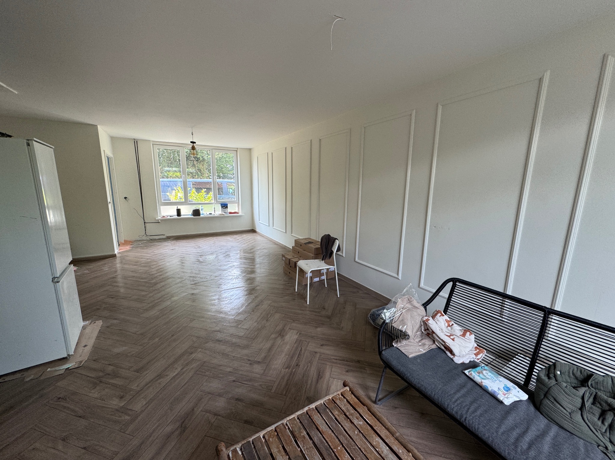 Pokój po remoncie z parkietem ułożonym w jodełkę, białe ściany z dekoracyjnymi listwami, meble tymczasowo ustawione w pomieszczeniu, widok na okno z zielenią za nim.