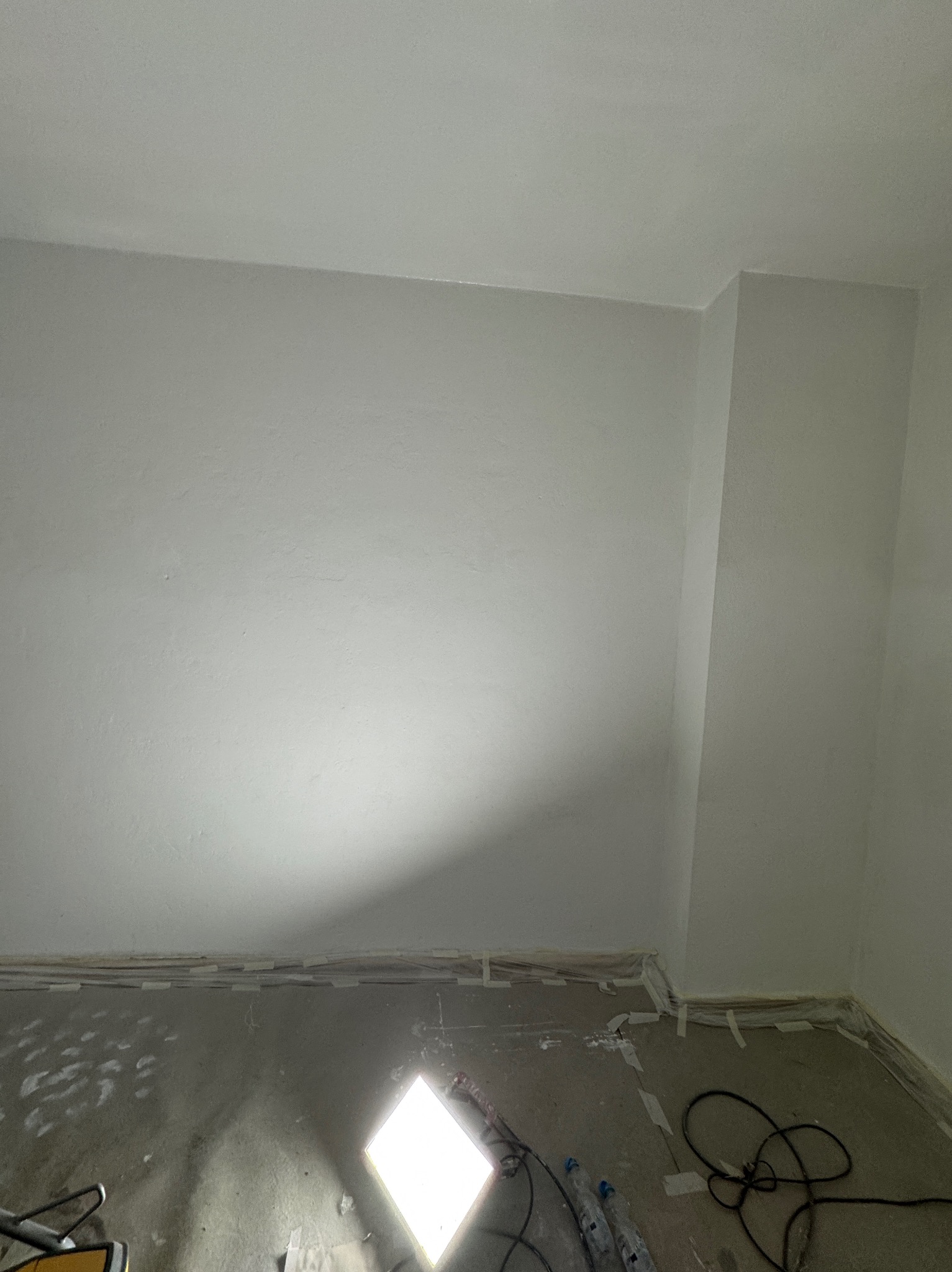 Świeżo pomalowane na biało ściany w pokoju, zabezpieczona taśmą malarską podłoga, lampa robocza na podłodze oświetla pomieszczenie