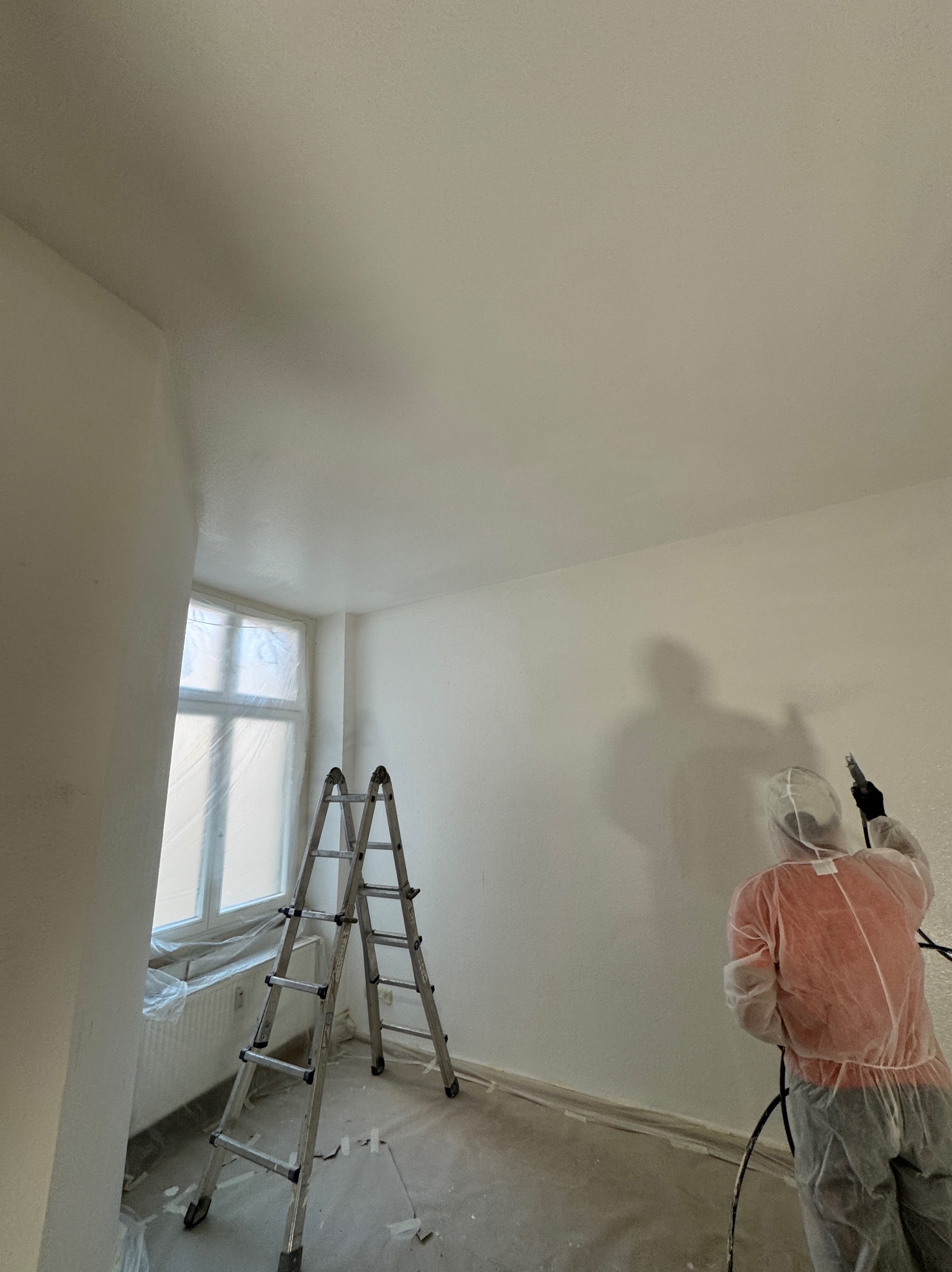Pracownik w kombinezonie ochronnym maluje ścianę natryskowo w pomieszczeniu z zabezpieczonym oknem i rozstawioną drabiną.