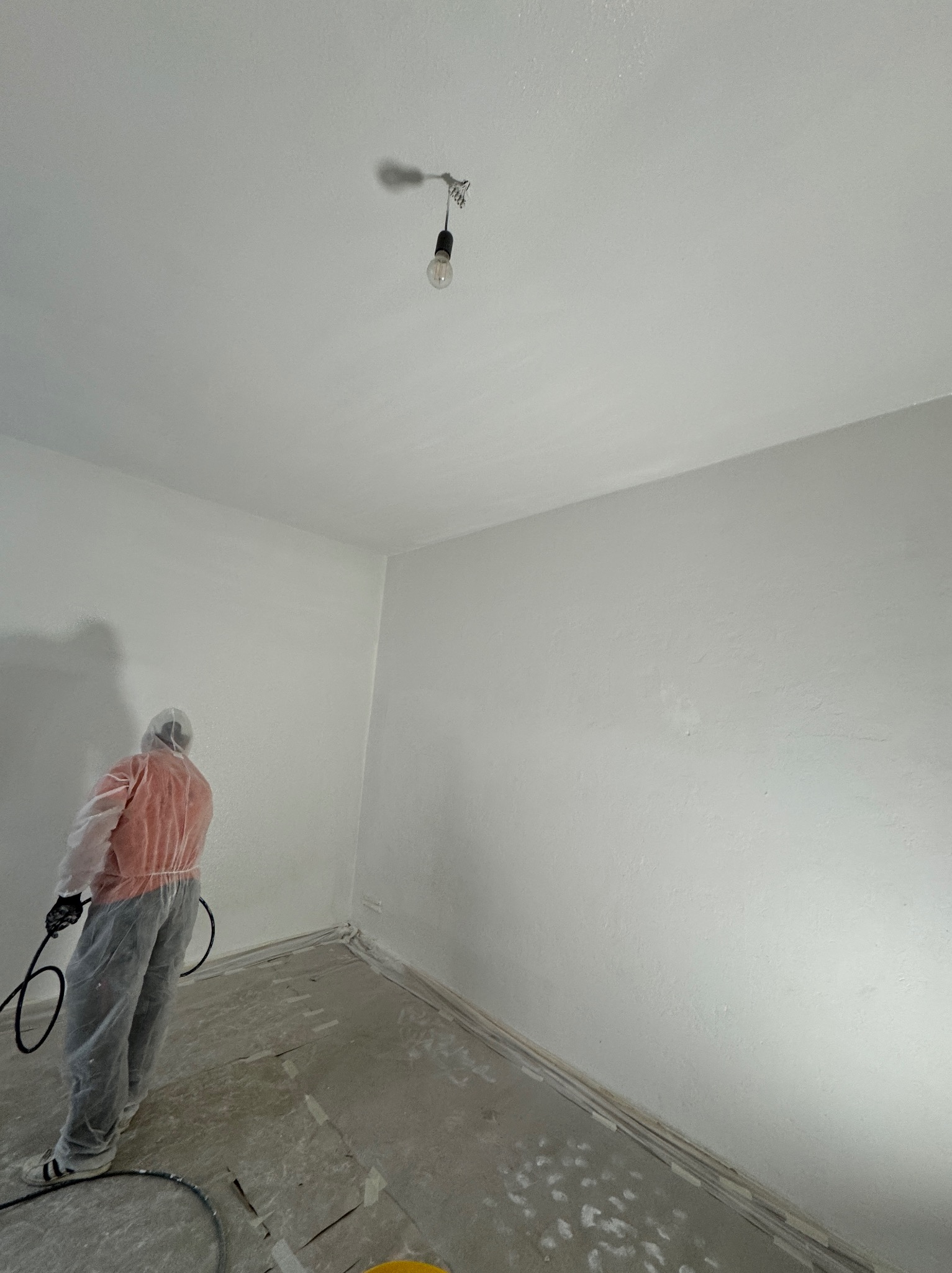 Pracownik w kombinezonie malarskim natryskuje białą farbę na ścianę w pokoju z gołą żarówką na suficie, widoczne ślady farby na podłodze zabezpieczonej folią.