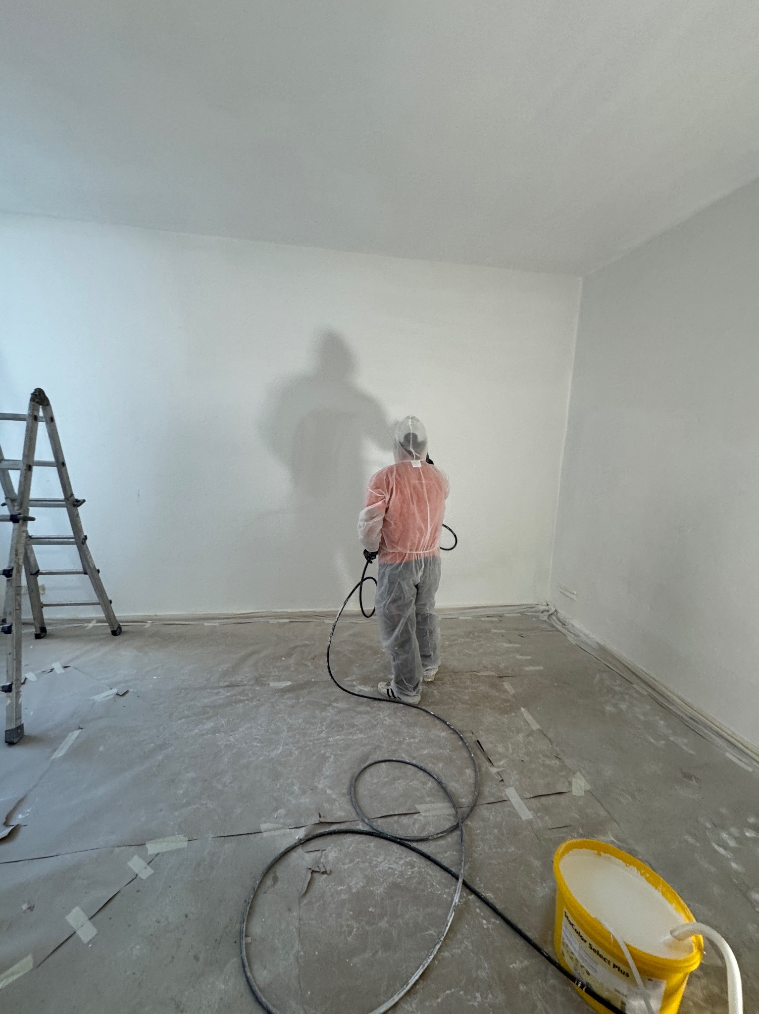 Pracownik w kombinezonie ochronnym maluje ściany pistoletem natryskowym w pomieszczeniu zabezpieczonym folią, widoczna drabina i wiadro z farbą.