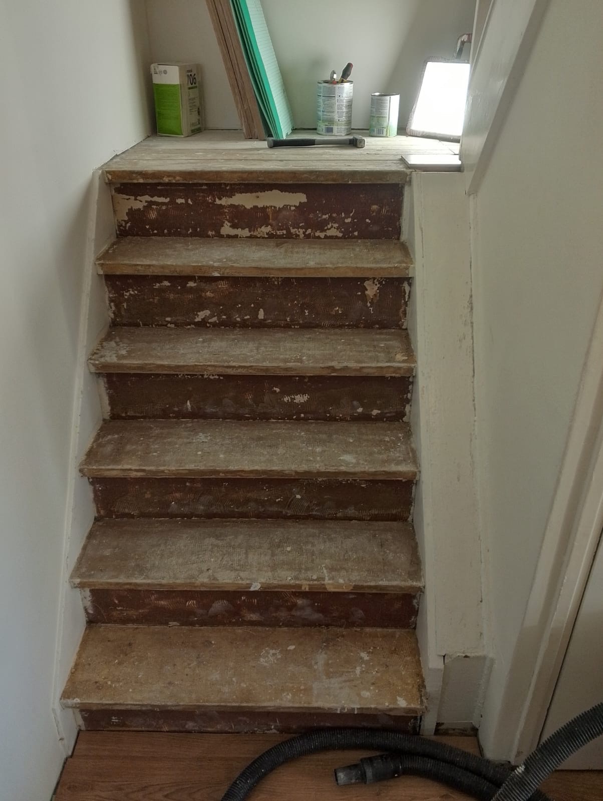 Schody drewniane w trakcie renowacji, z widocznymi śladami starej farby i przygotowaniem do malowania, narzędzia malarskie na górnym podeście, odkurzacz u dołu schodów.