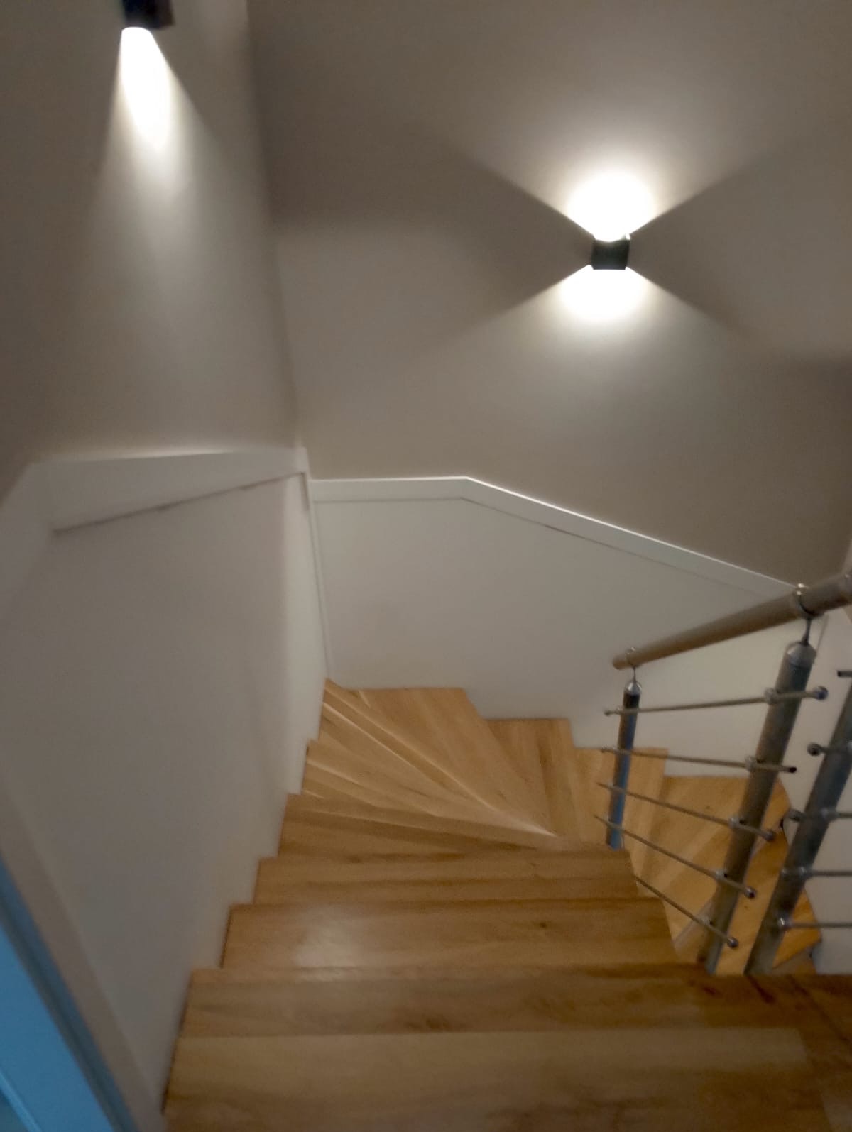 Drewniane schody spiralne z metalową balustradą, oświetlone dwoma nowoczesnymi lampami ściennymi, widok z góry.