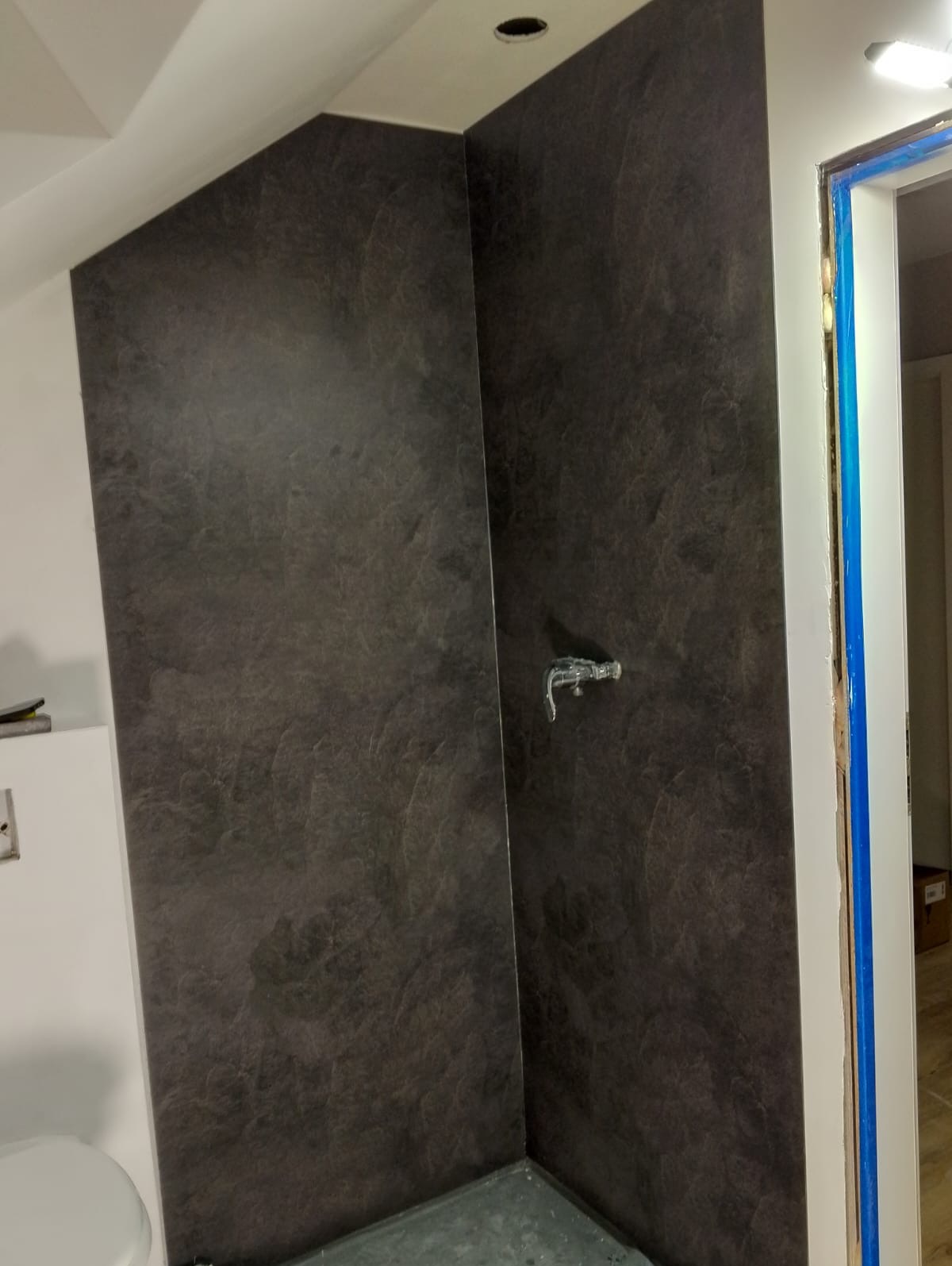 Ściana kabiny prysznicowej wykończona ciemną, strukturalną okładziną imitującą kamień, z zamontowaną baterią prysznicową, widoczna fragment toalety oraz zabezpieczone ościeże drzwi.