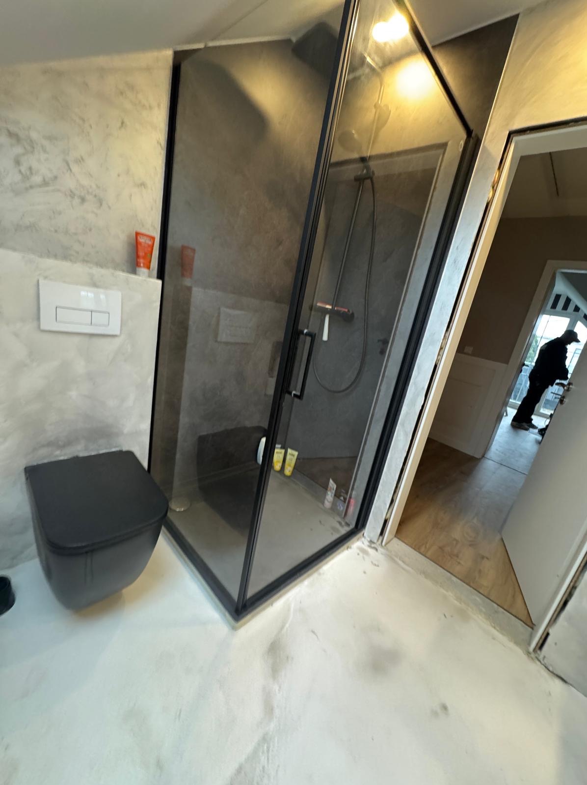 Nowoczesna łazienka z czarną armaturą: kabina prysznicowa walk-in z grafitowymi płytkami, wisząca toaleta z ukrytym systemem spłukiwania i biała posadzka z żywicy epoksydowej. W tle widoczna osoba...