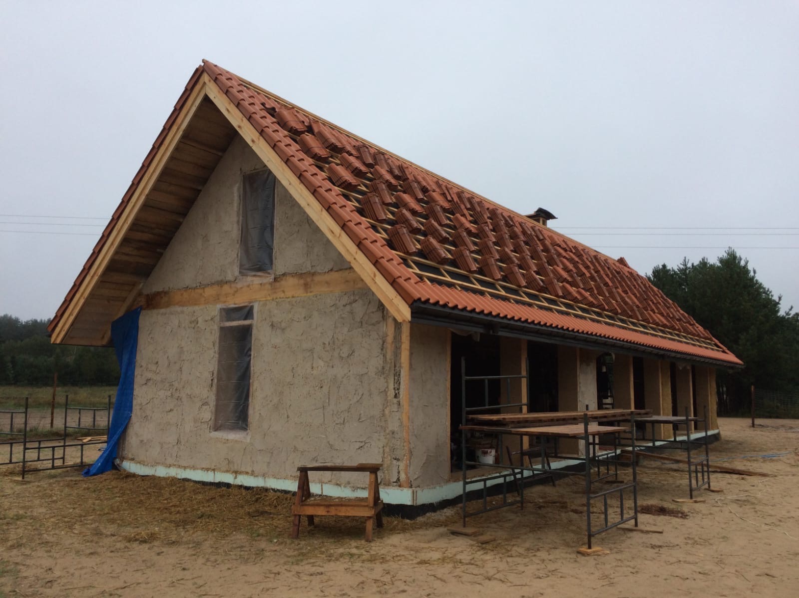 Budowa domu z gliny z częściowo ułożoną czerwoną dachówką, okna zabezpieczone folią, widoczne rusztowania i konstrukcja drewniana dachu.