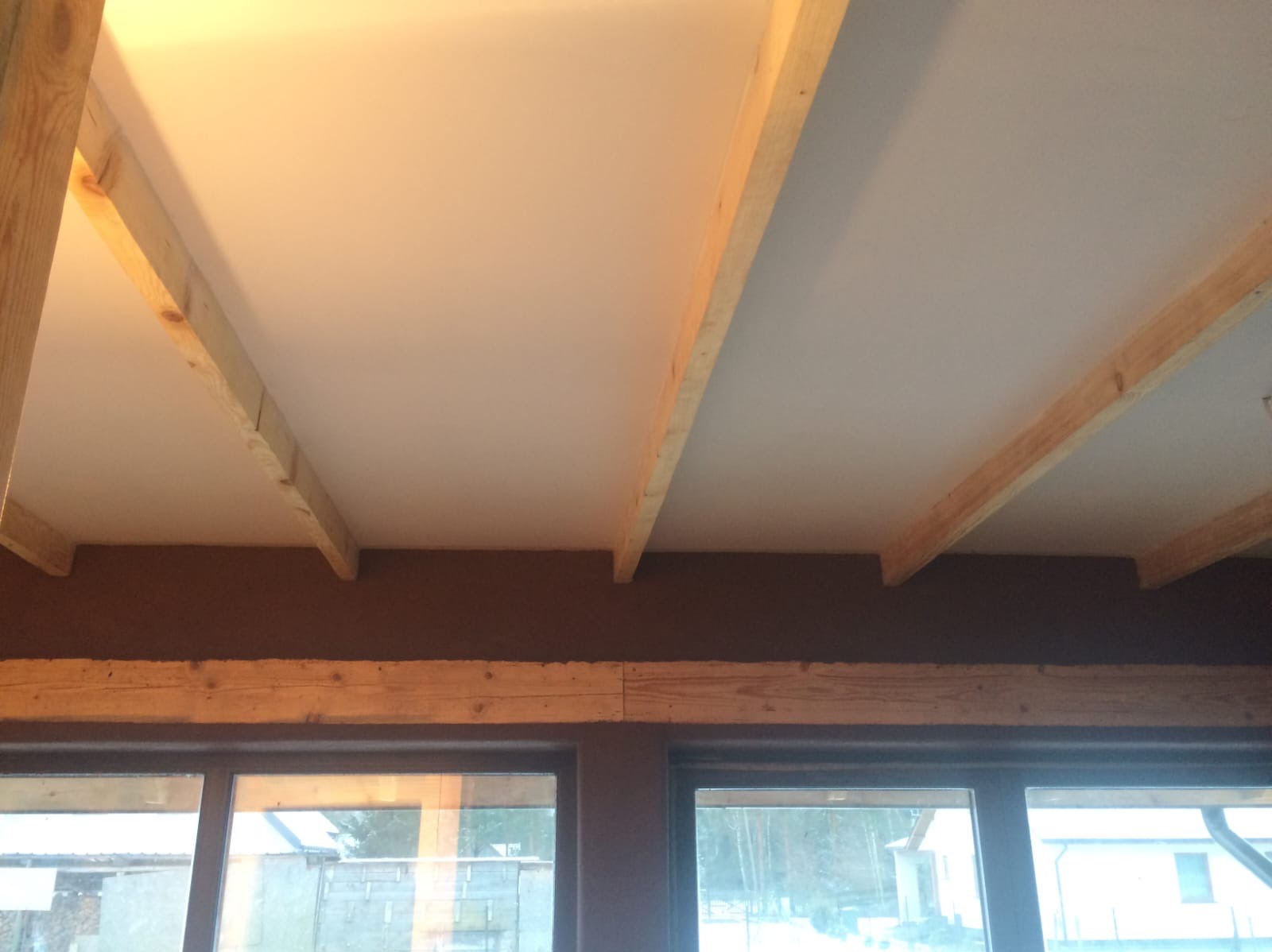 Biały sufit z drewnianymi belkami konstrukcyjnymi w nowym domu, widok z dołu na okna i zimowy krajobraz za nimi.