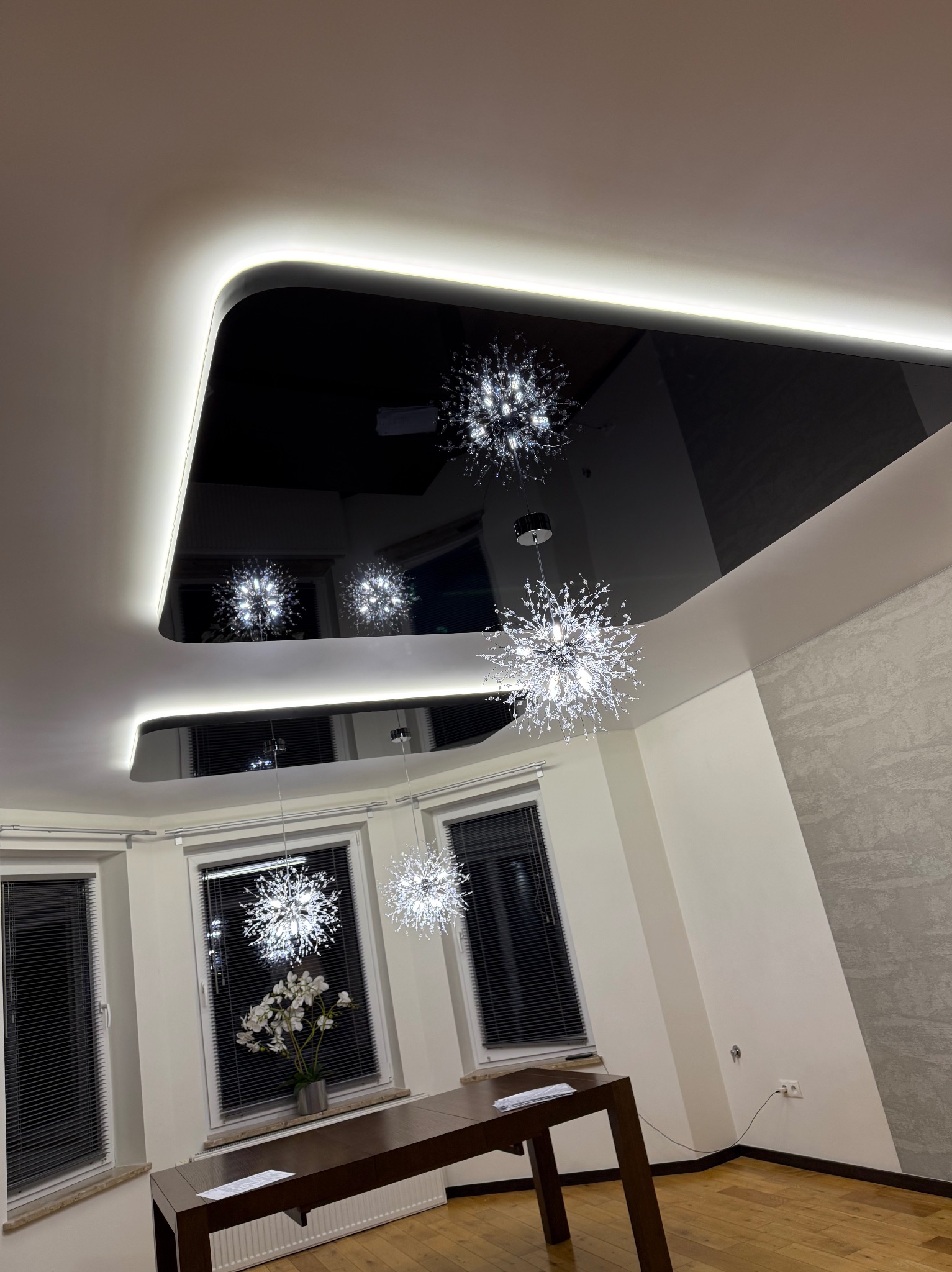 Nowoczesny czarny sufit napinany z podświetleniem LED i odbijającymi się w nim trzema kryształowymi żyrandolami w jasnym pomieszczeniu z drewnianą podłogą i stołem.