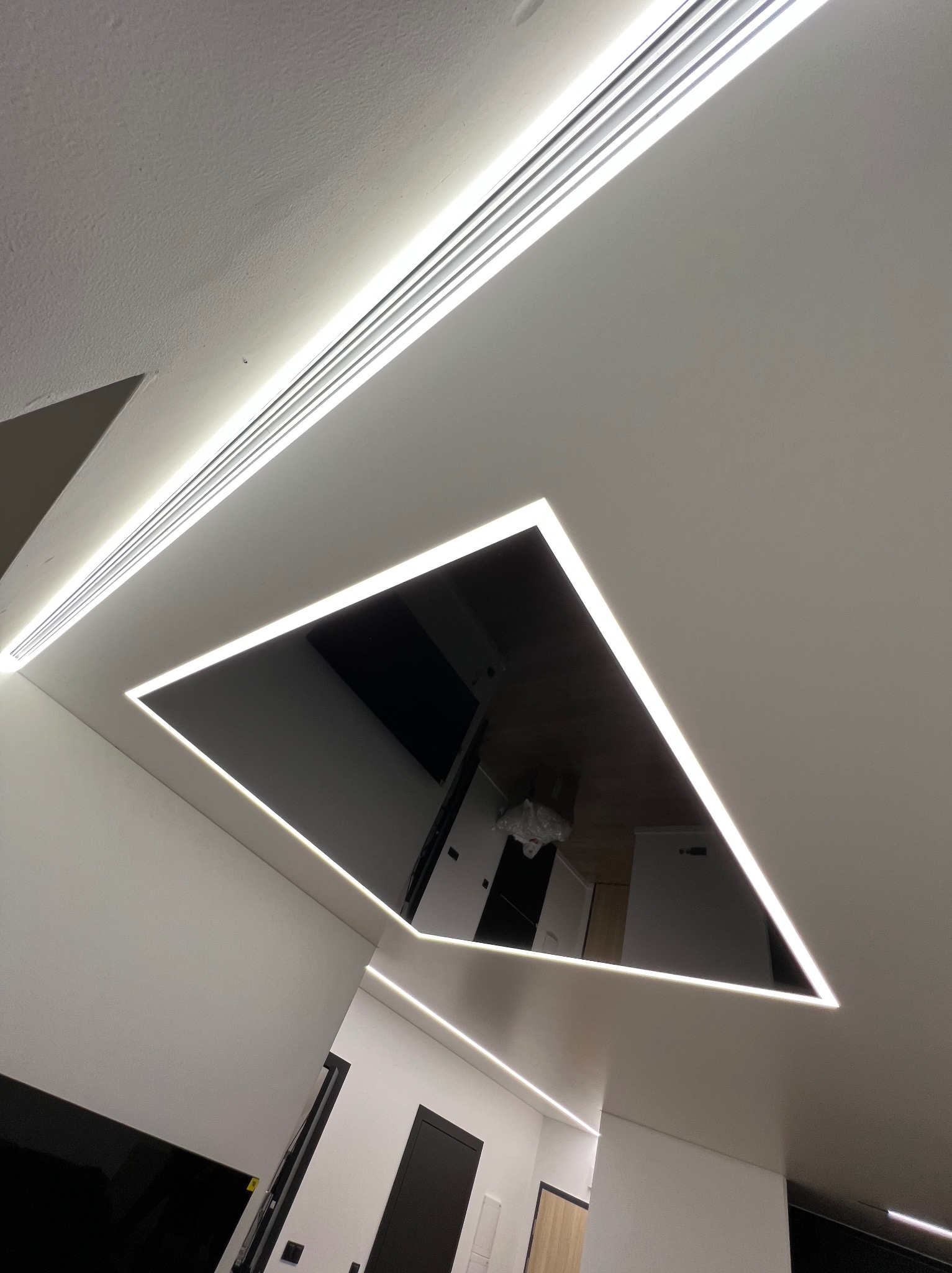 Nowoczesny sufit napinany z czarnym lustrzanym panelem w kształcie trójkąta, obramowanym białym oświetleniem LED, z widocznym odbiciem wnętrza pomieszczenia.