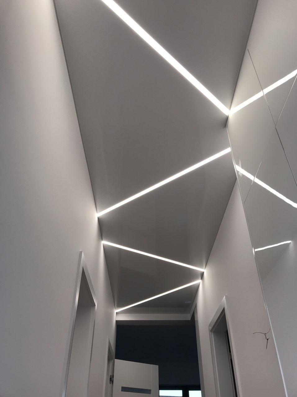 Biały sufit z liniowym oświetleniem LED tworzącym geometryczny wzór w korytarzu, widoczne drzwi i fragment ściany z połyskiem.