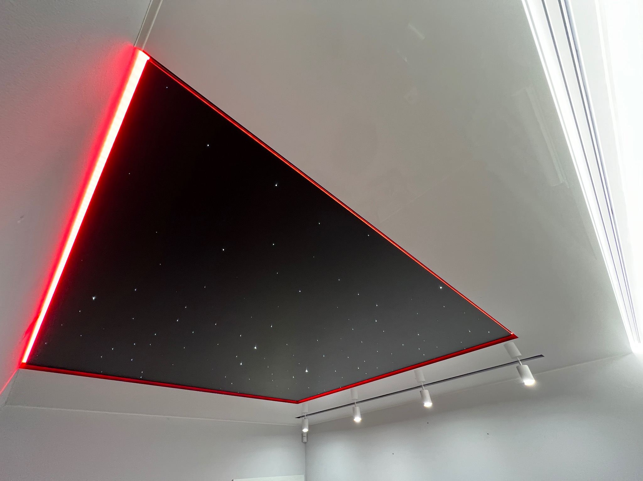 Czarny, trójkątny fragment sufitu z efektem gwiazd, obramowany czerwoną listwą LED, wkomponowany w biały, błyszczący sufit. Widoczne reflektory punktowe.