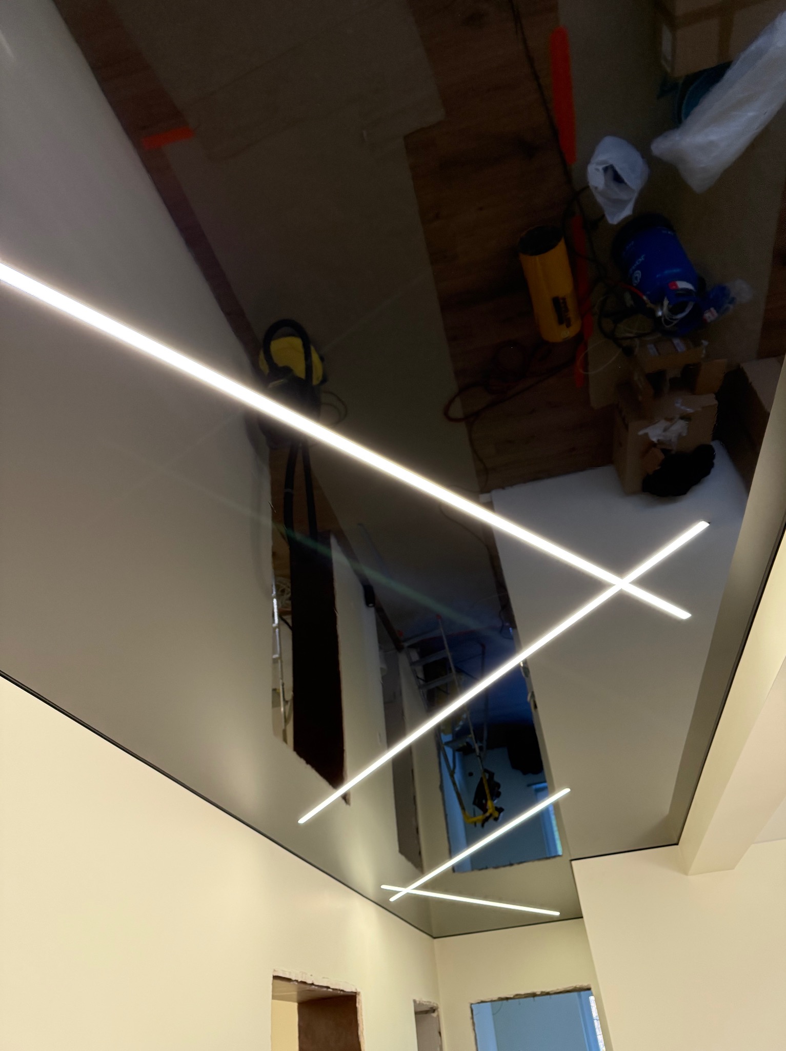 Błyszczący, czarny sufit z listwami LED tworzącymi geometryczny wzór, widoczne odbicia narzędzi i materiałów budowlanych sugerujące trwający montaż.