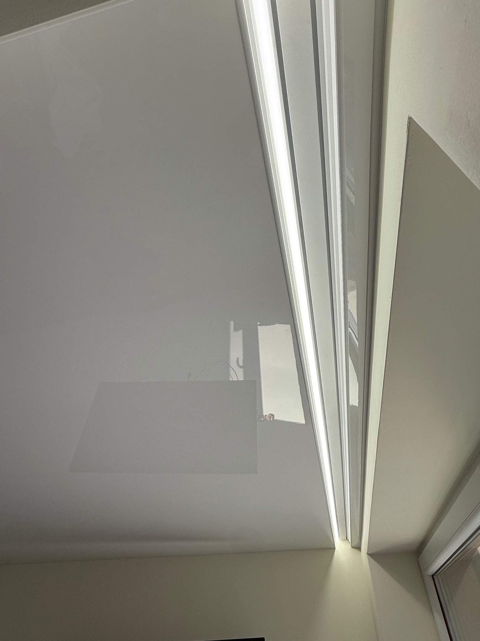 Biały, błyszczący sufit z listwami LED, widoczny fragment okna i ściany, zarys instalacji elektrycznej na suficie.
