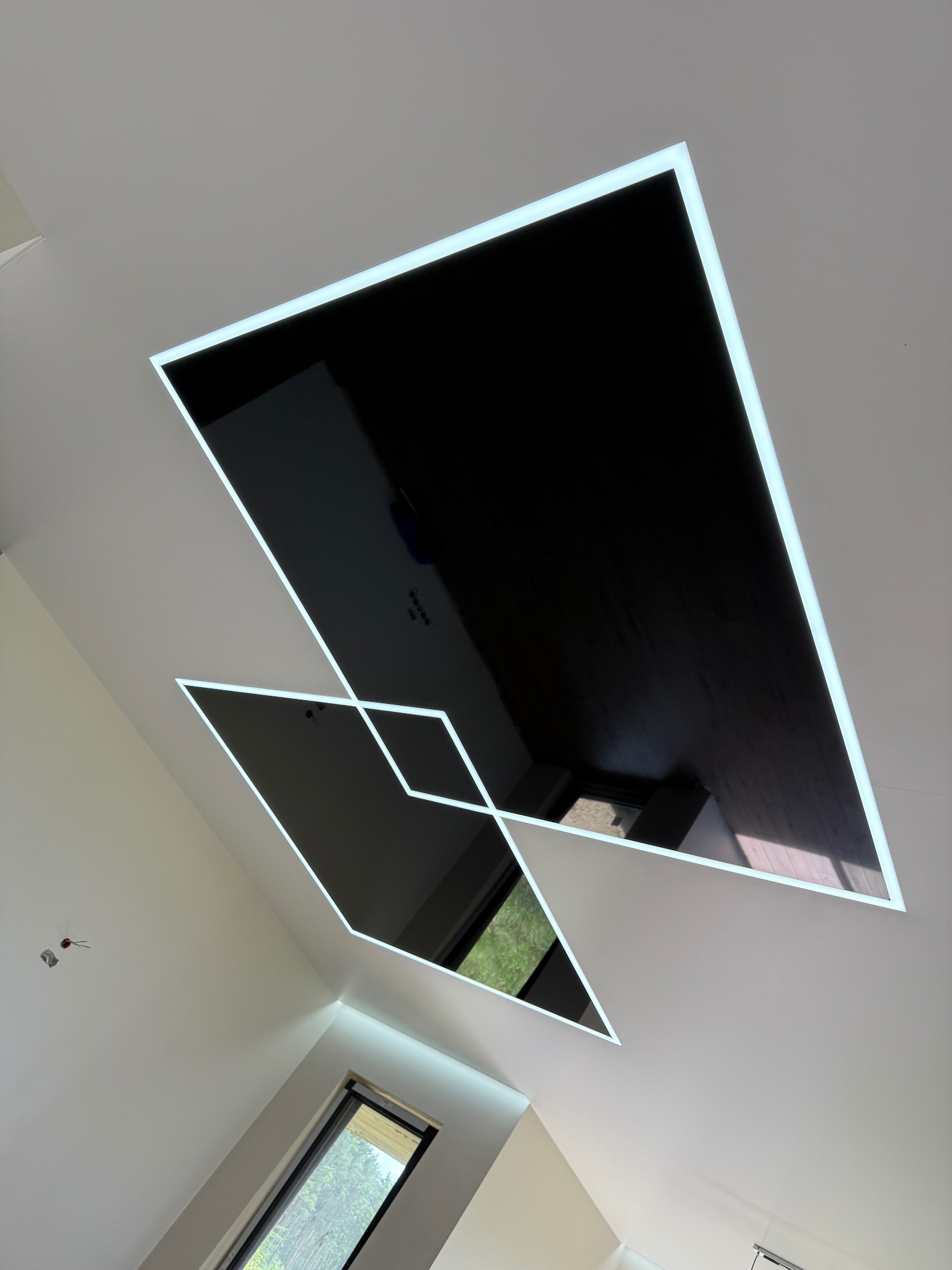 Nowoczesny sufit z czarnymi panelami i podświetleniem LED w kształcie geometrycznym, odbijający światło okna. Minimalistyczny design wnętrza.