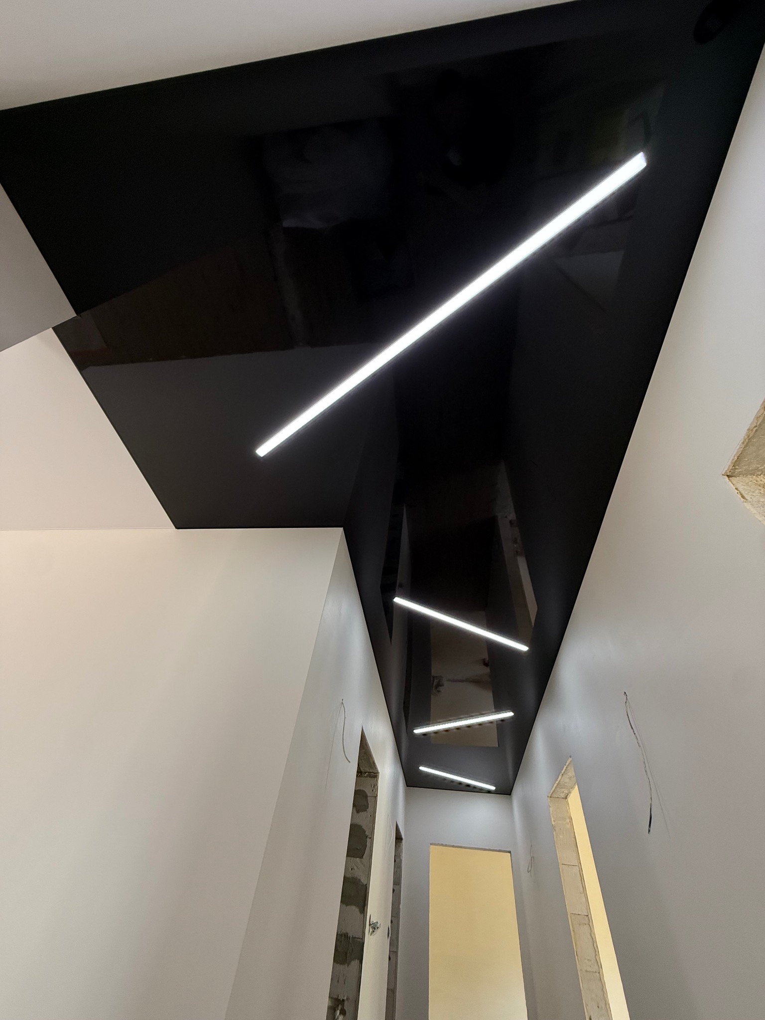 Nowoczesny, czarny, błyszczący sufit z liniowym oświetleniem LED w korytarzu, widziany z dołu. Białe ściany, surowy stan wykończenia.