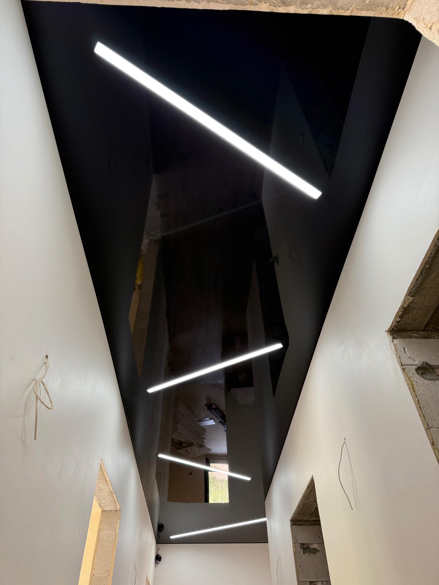 Czarny sufit napinany z nowoczesnym oświetleniem LED w kształcie linii, odbijający wnętrze w wykończonym pomieszczeniu w trakcie remontu.