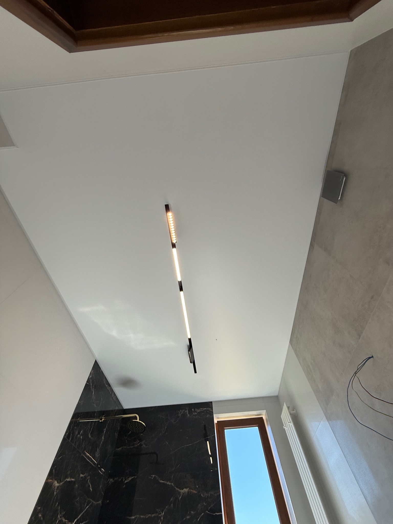Biały sufit w łazience z nowoczesnym, podłużnym oświetleniem LED. Widoczne elementy wykończenia: marmurowe płytki i czarna ściana prysznica.