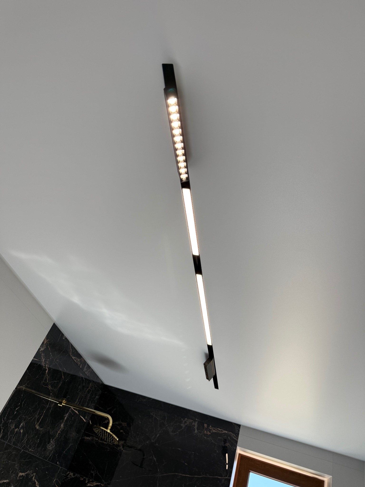 Biały sufit z czarną, podłużną lampą LED w łazience z czarnymi marmurowymi płytkami i złotą armaturą. Minimalistyczny design, nowoczesne oświetlenie.