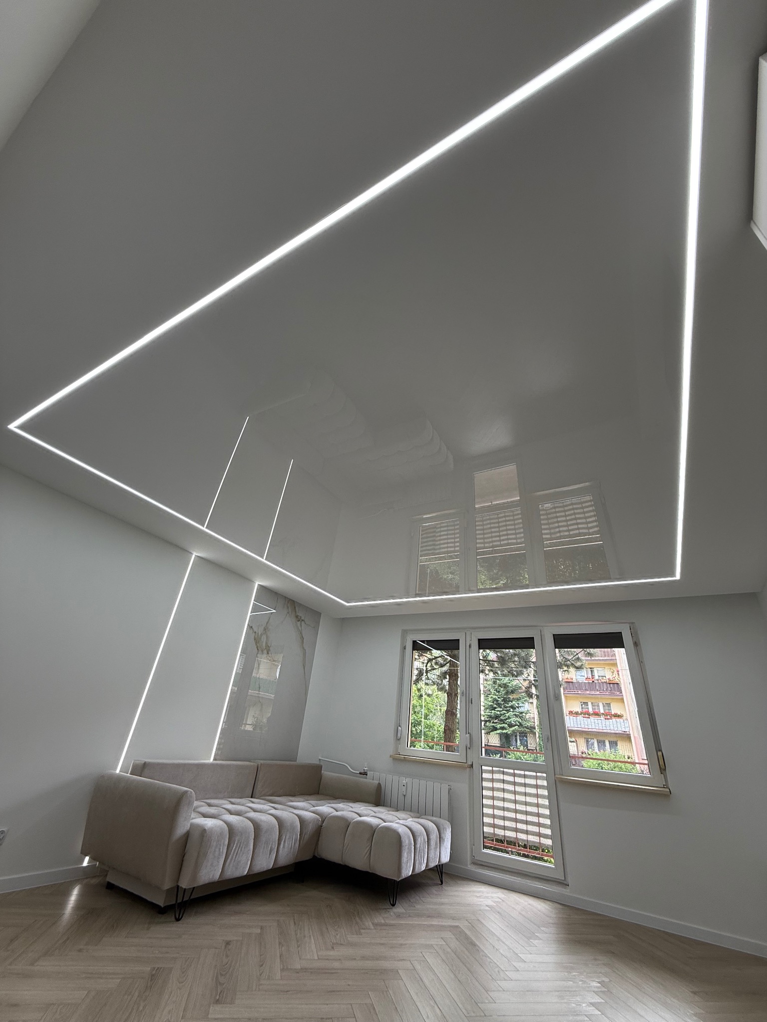 Biały, błyszczący sufit z podświetleniem LED w kształcie prostokąta i linii, odbijający światło z okna w nowoczesnym salonie z beżową sofą narożną.
