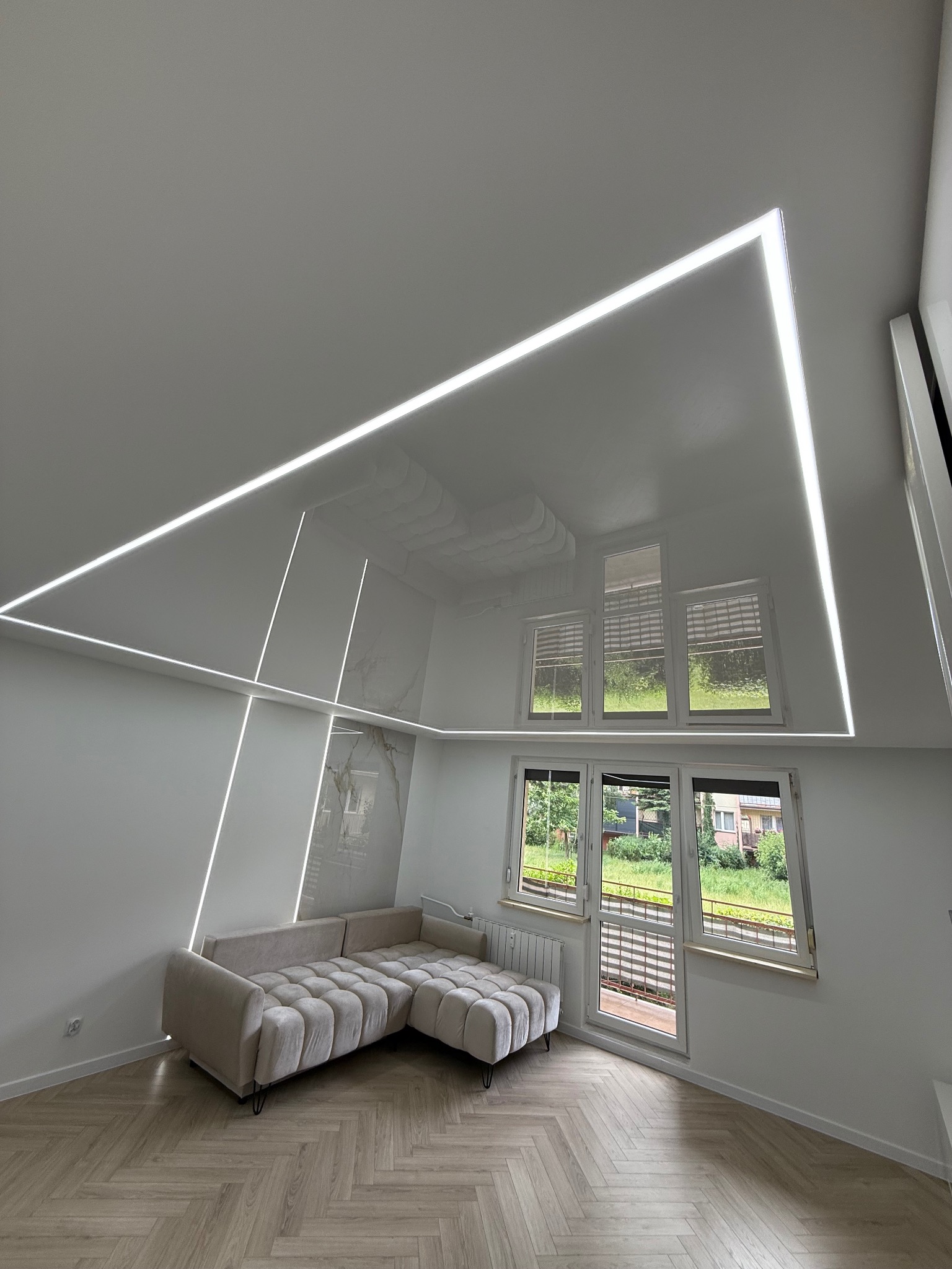 Nowoczesny salon z połyskującym sufitem i oświetleniem LED w Warszawie. Widoczny narożnik i okna z widokiem na zieleń. Minimalistyczny design.