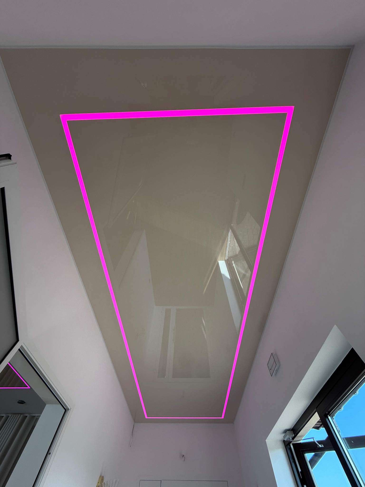 Błyszczący, beżowy sufit z wbudowanym oświetleniem LED w kolorze fuksji, tworzącym geometryczny kształt. Widok z dołu, okno w tle.