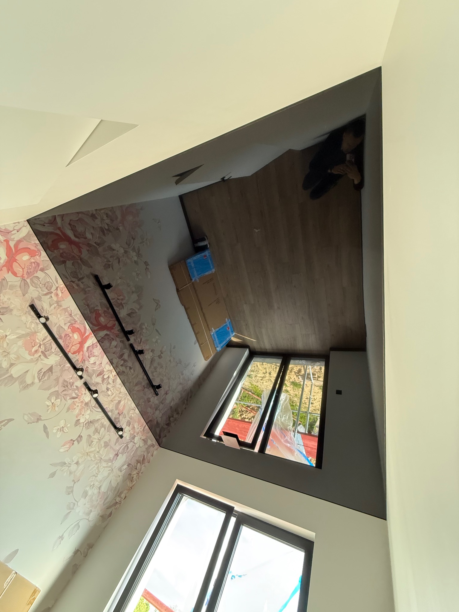 Nowoczesne wnętrze z oknami, wzorzystą tapetą i elementami sufitu podwieszanego. Widoczne reflektory sufitowe i kartony, prawdopodobnie z materiałami wykończeniowymi.