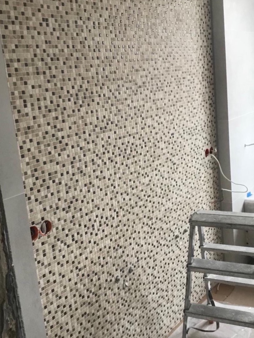 Ściana w łazience wyłożona mozaiką w odcieniach beżu i brązu, widoczne wystające rury i drabina. Efektowna kompozycja małych płytek tworzy unikalny wzór.