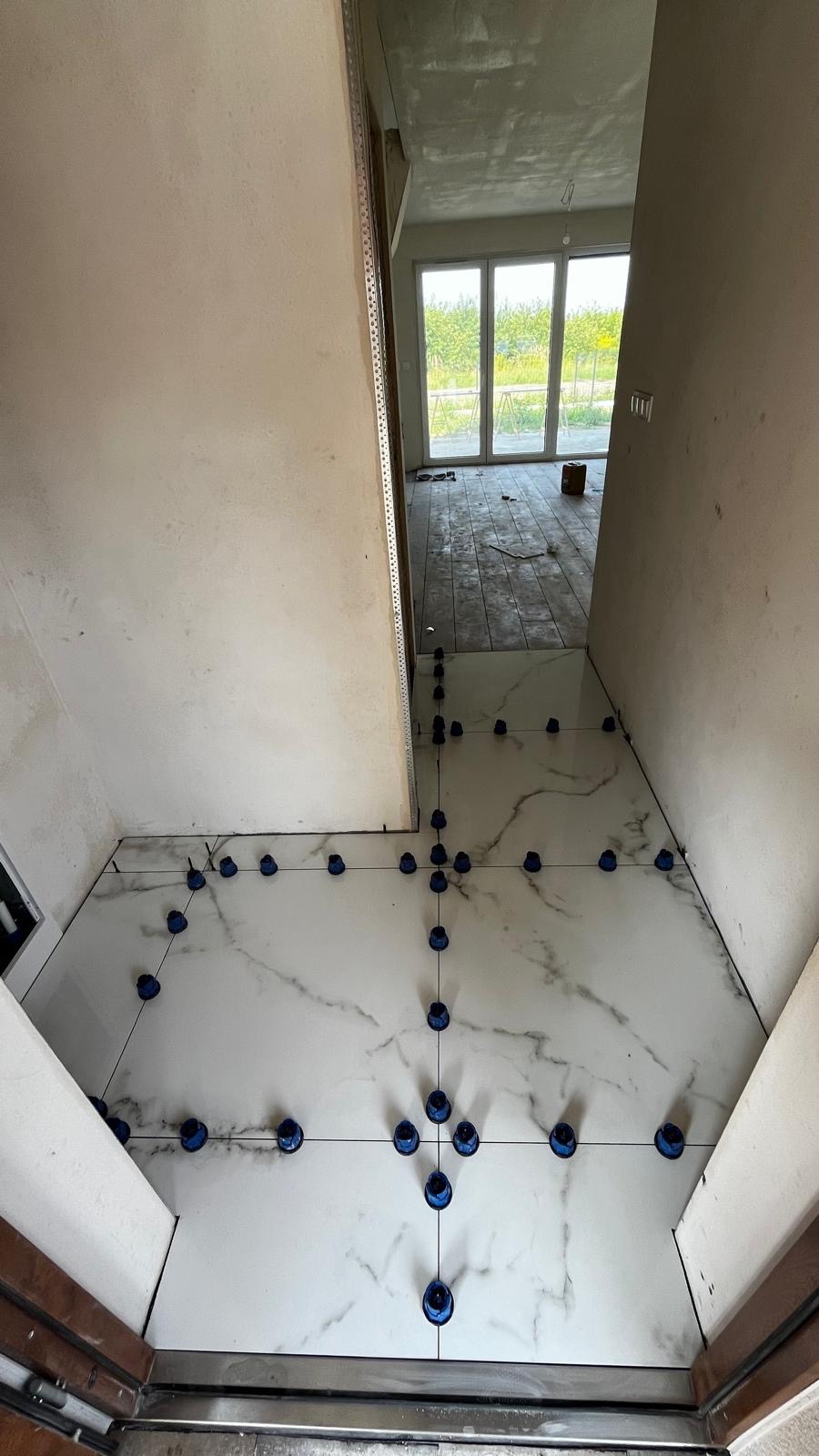 Układanie białych płytek imitujących marmur z użyciem niebieskich klipsów poziomujących w korytarzu. W tle widoczne okno i surowy stan wykończenia.