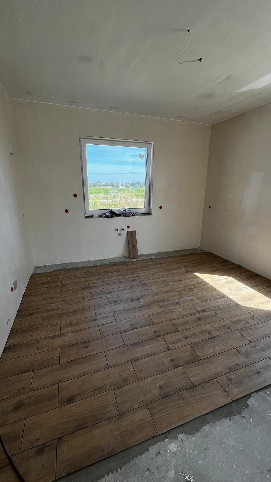 Układanie podłogi z płytek imitujących drewno w jasnym pokoju z oknem. Widoczne niedokończone ściany i wystające kable elektryczne.