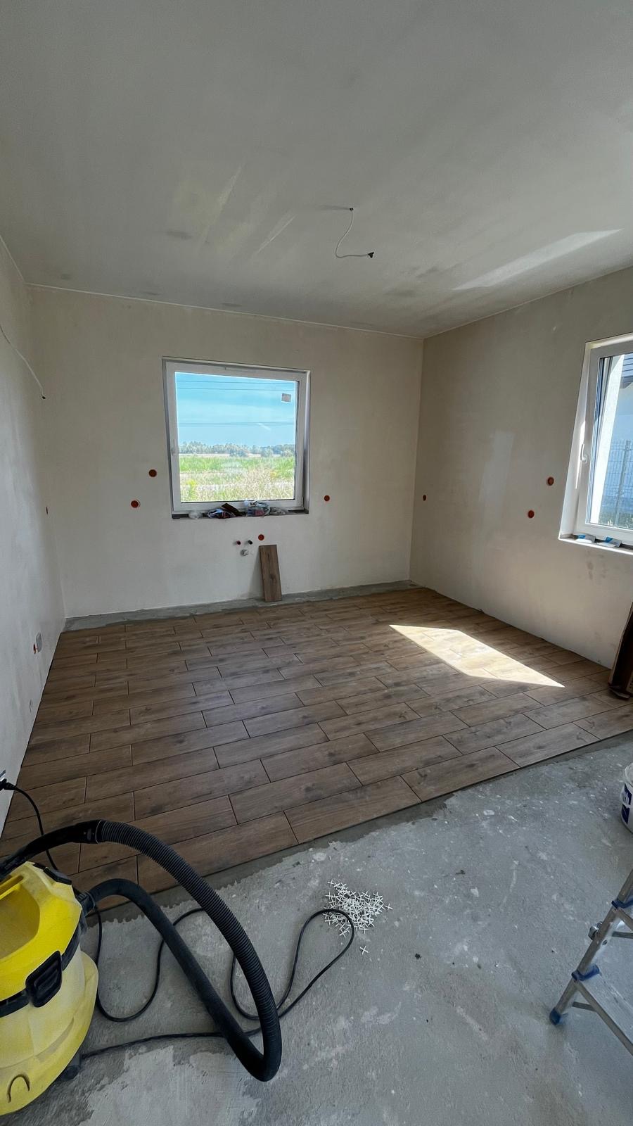 Układanie brązowych płytek imitujących drewno w jasnym pokoju. Widoczny odkurzacz przemysłowy i krzyżyki dystansowe. Częściowo wykończone ściany.