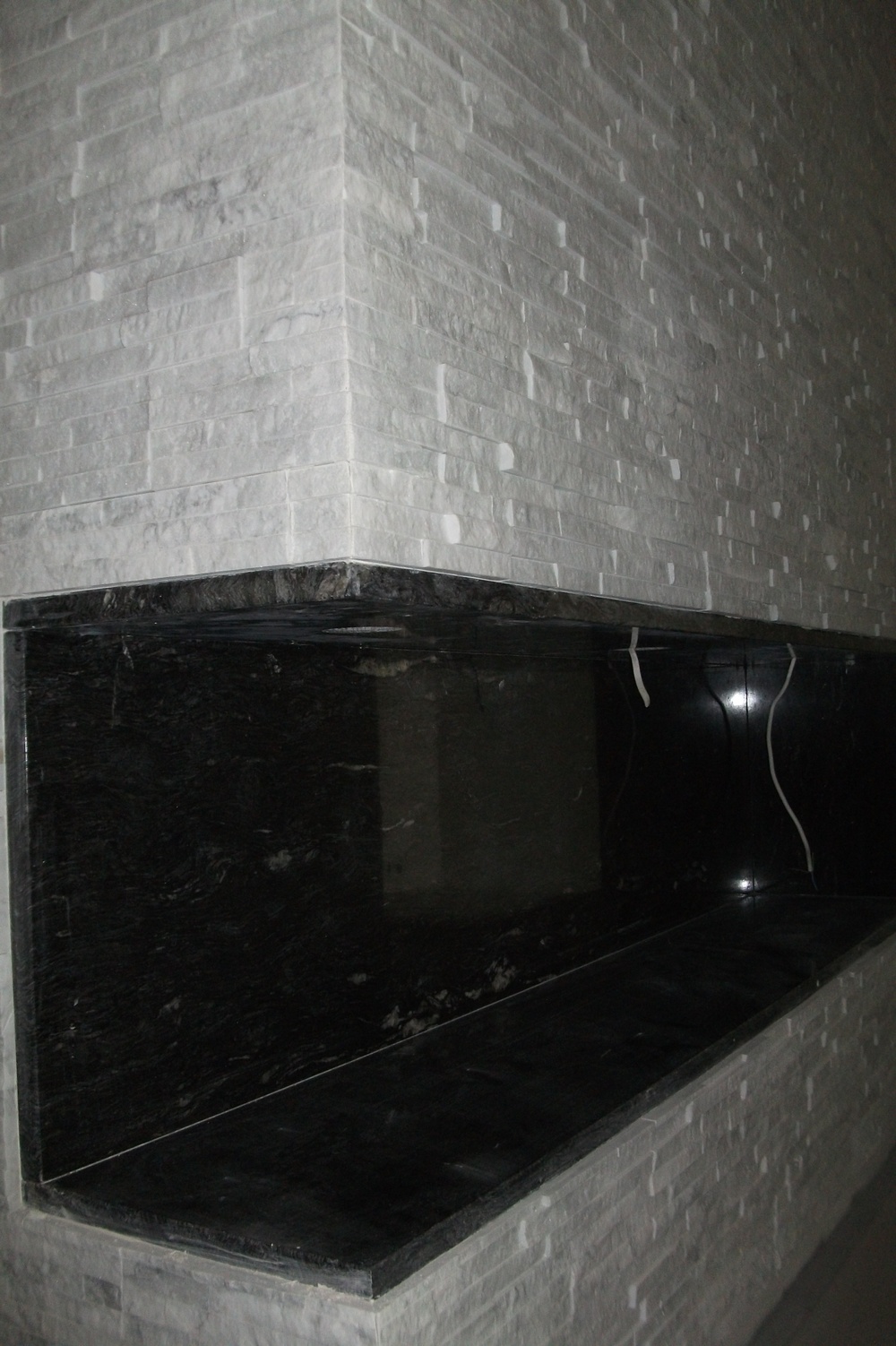 Ściana wykończona dekoracyjną płytką imitującą kamień, z wnęką wyłożoną czarnym marmurem. Widoczne przewody elektryczne we wnęce.