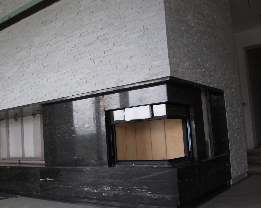 Nowoczesny kominek z obudową z czarnego kamienia i ściana wykończona płytkami imitującymi kamień w odcieniach szarości, widoczne elementy wkładu kominkowego.