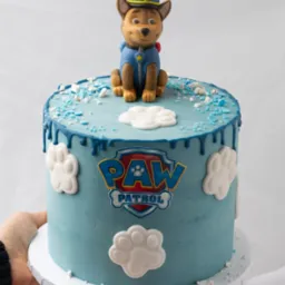 Tort urodzinowy w stylu Psi Patrol, z figurką Chase'a na wierzchu, błękitnym lukrem, białymi dekoracjami łapek i napisem 'Paw Patrol', trzymany przez osobę na białym tle.