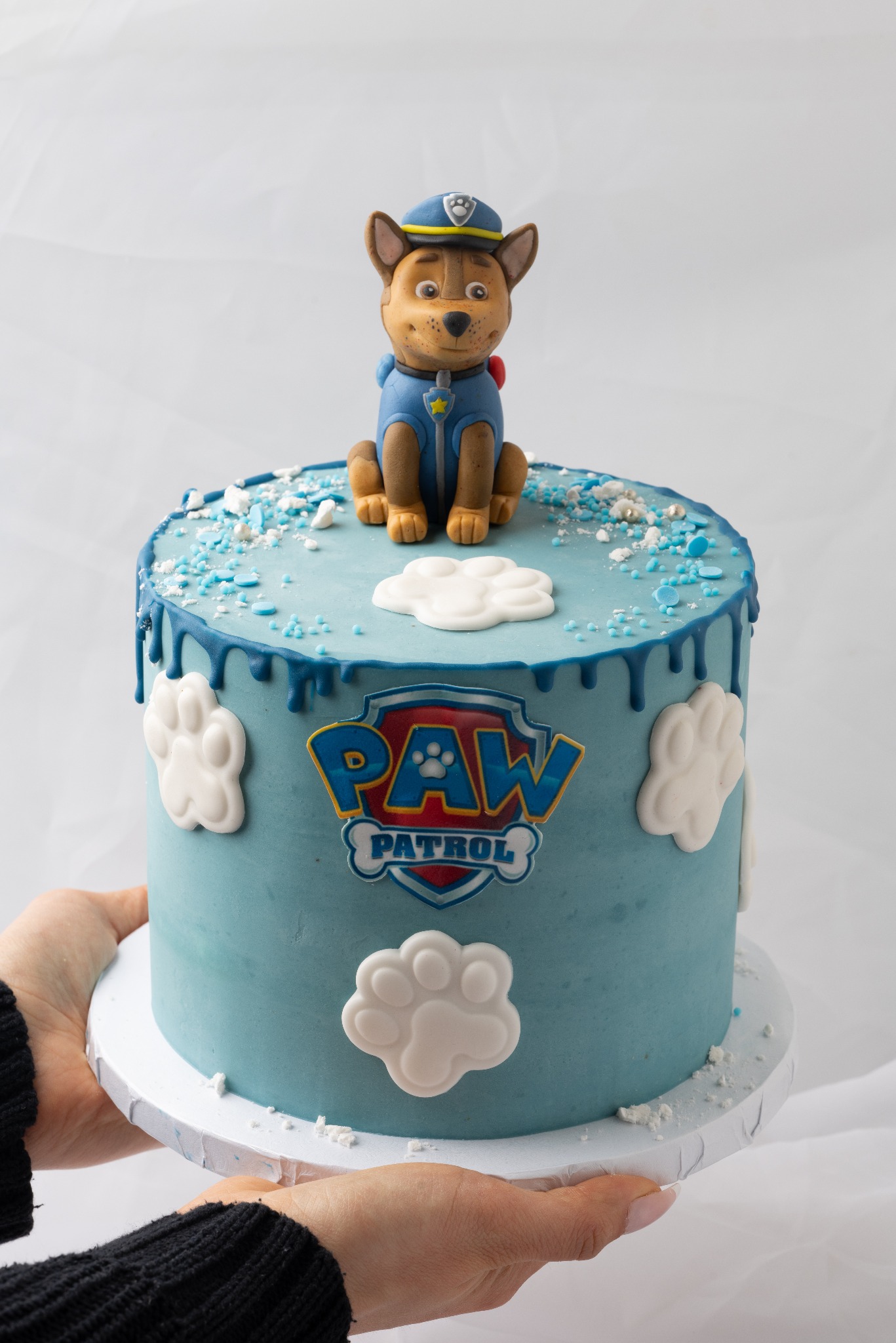 Tort urodzinowy w stylu Psi Patrol, z figurką Chase'a na wierzchu, błękitnym lukrem, białymi dekoracjami łapek i napisem 'Paw Patrol', trzymany przez osobę na białym tle.