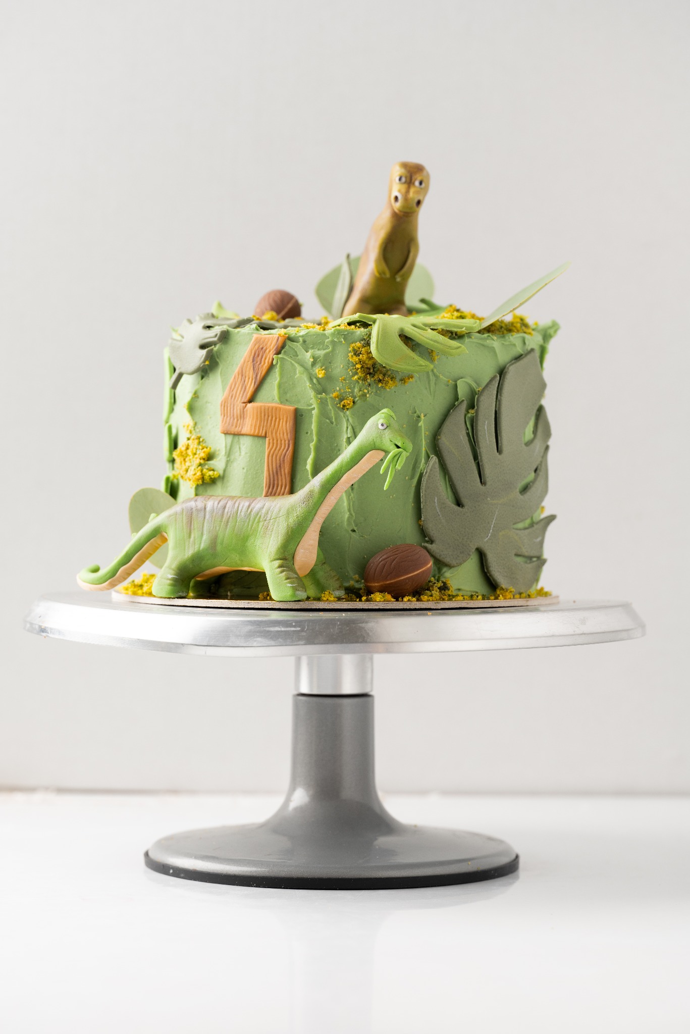 Tort urodzinowy z zielonym kremem i dekoracjami z masy cukrowej w kształcie dinozaurów i liści na srebrnej paterze.