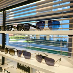 CENTRUM ZDROWIA OKA Michał Nowak - Ekspozycja okularów korekcyjnych i przeciwsłonecznych różnych marek na białych półkach sklepowych, z widokiem na budynki przez żaluzje w tle.