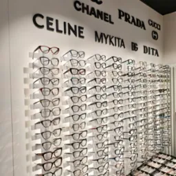 Ekspozycja oprawek okularów w salonie optycznym, marki takie jak Dior, Chanel, Prada, Celine, Mykita i Dita na ścianie, różnorodne modele w rzędach i w gablotach.