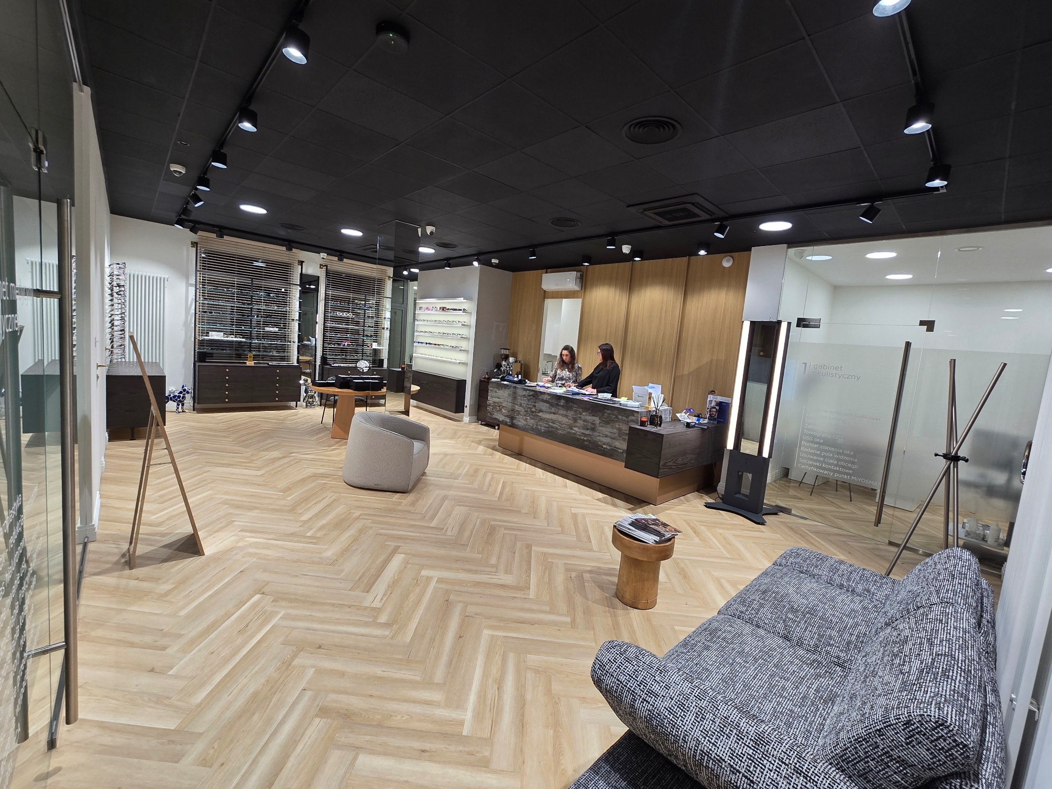 Przestronne wnętrze salonu optycznego z drewnianą podłogą w jodełkę, recepcją z dwoma pracownikami, ekspozycją oprawek okularów i wygodnymi fotelami dla klientów, oświetlone punktowymi lampami...