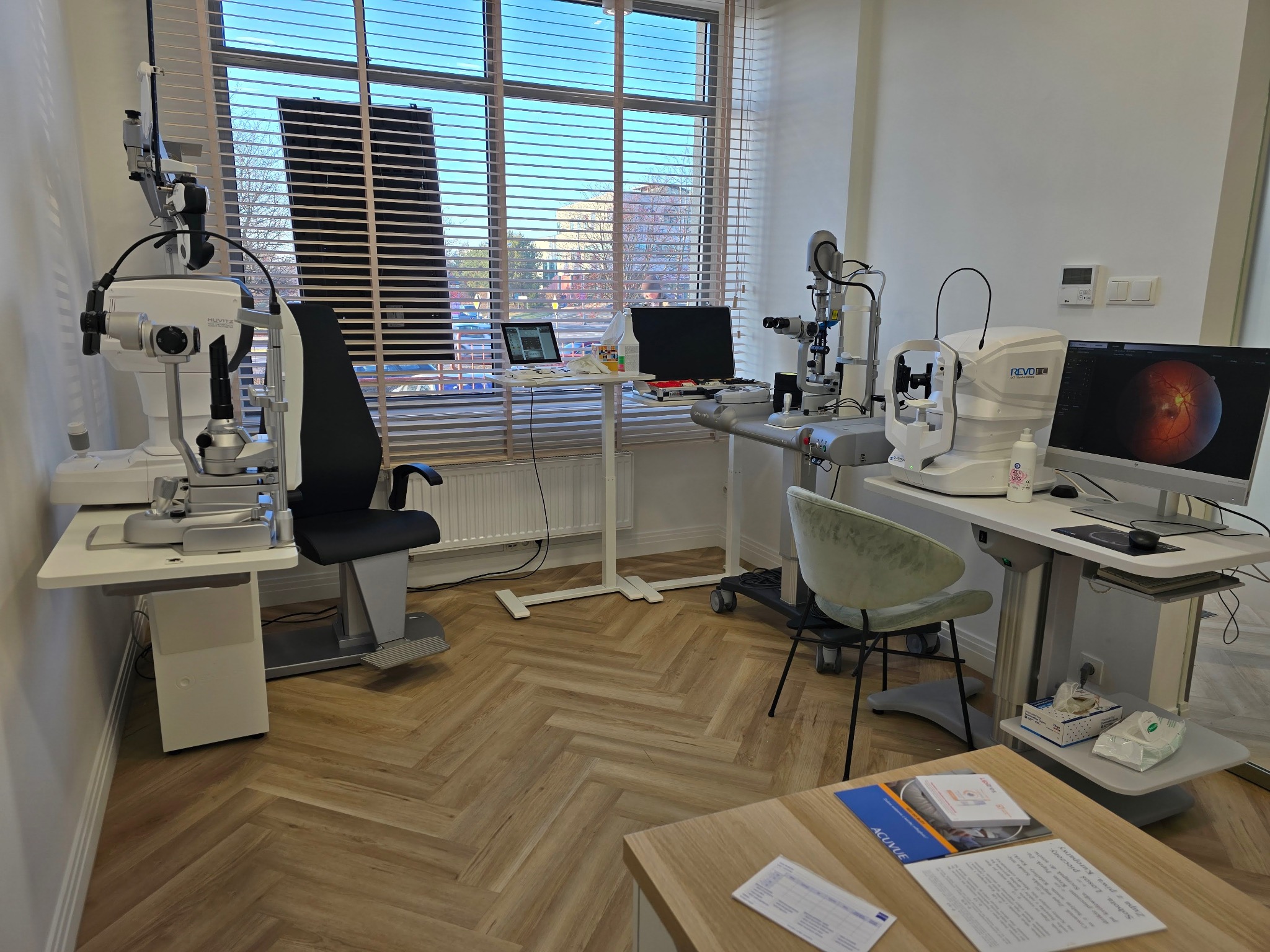 Jasne wnętrze gabinetu lekarskiego z dwoma stanowiskami do badania wzroku, wyposażonymi w specjalistyczny sprzęt diagnostyczny, monitory i krzesła. W tle widoczne okno z żaluzjami i budynek...