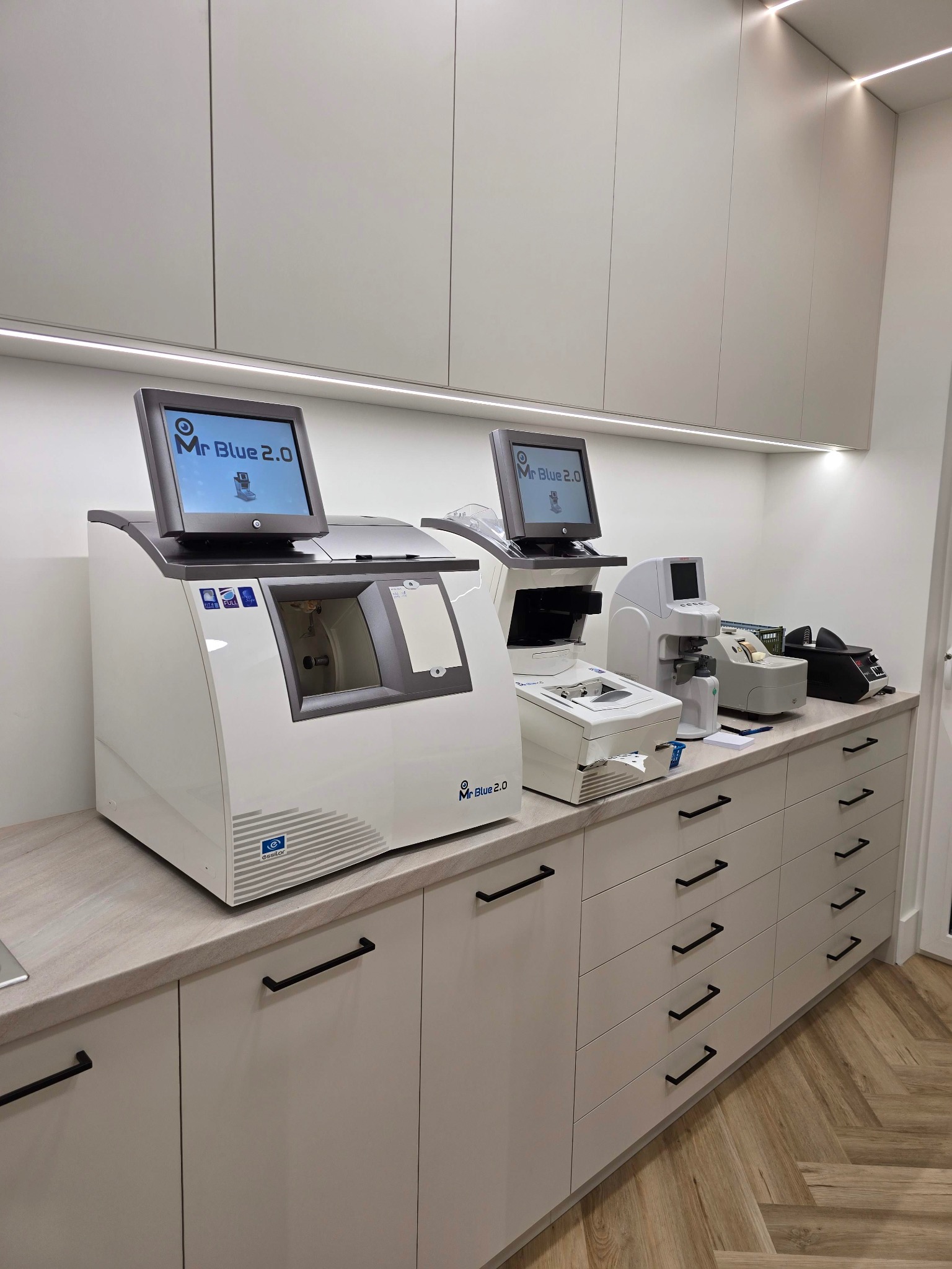 Nowoczesne gabinetowe wyposażenie: Dwa automatyczne szlifierki do soczewek okularowych, model Mr Blue 2.0, stoją na blacie przy ścianie z szafkami i oświetleniem LED.