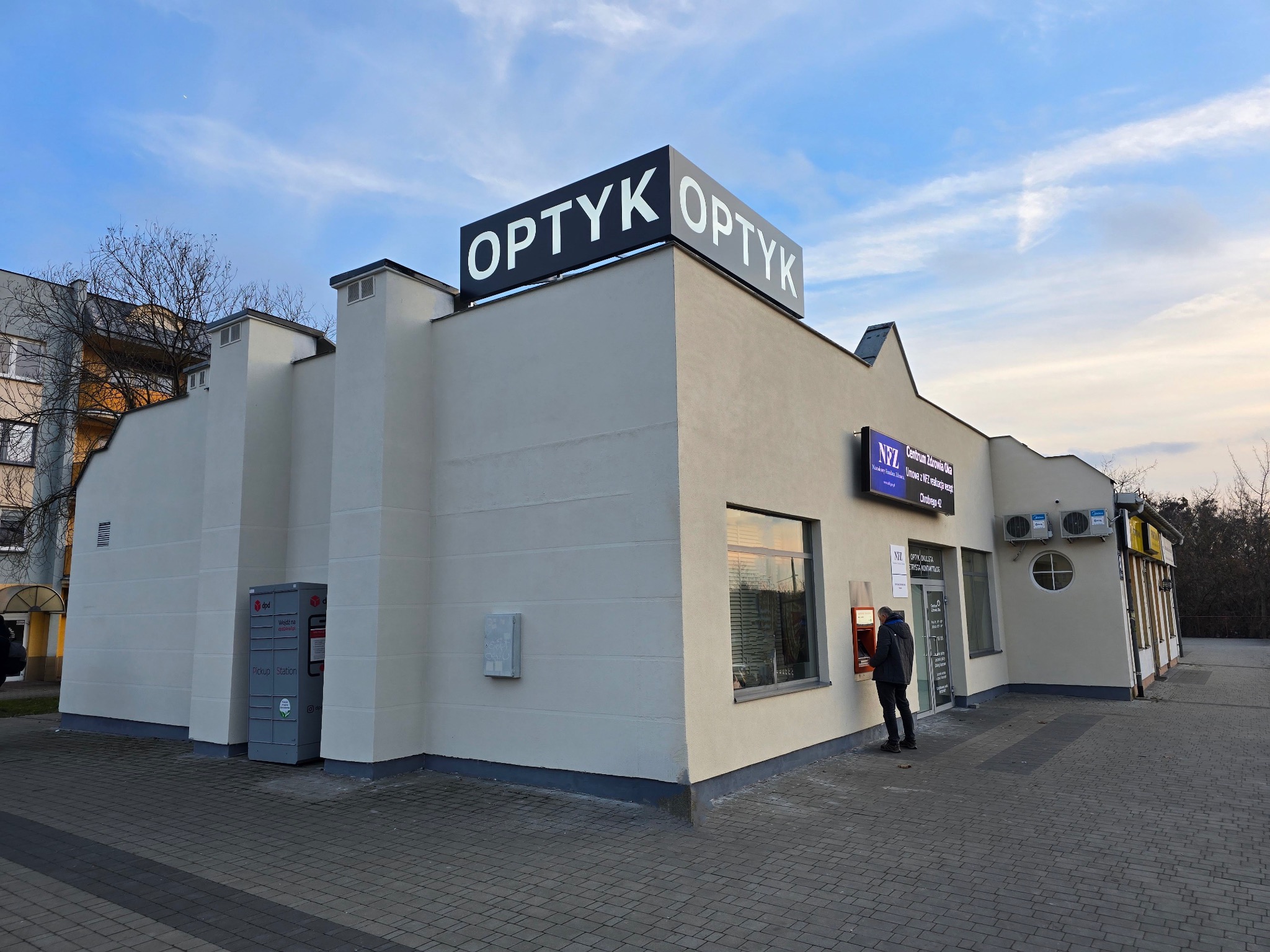 Budynek z szyldem 'OPTYK' na dachu i paczkomatem obok, klient przy bankomacie przy wejściu.
