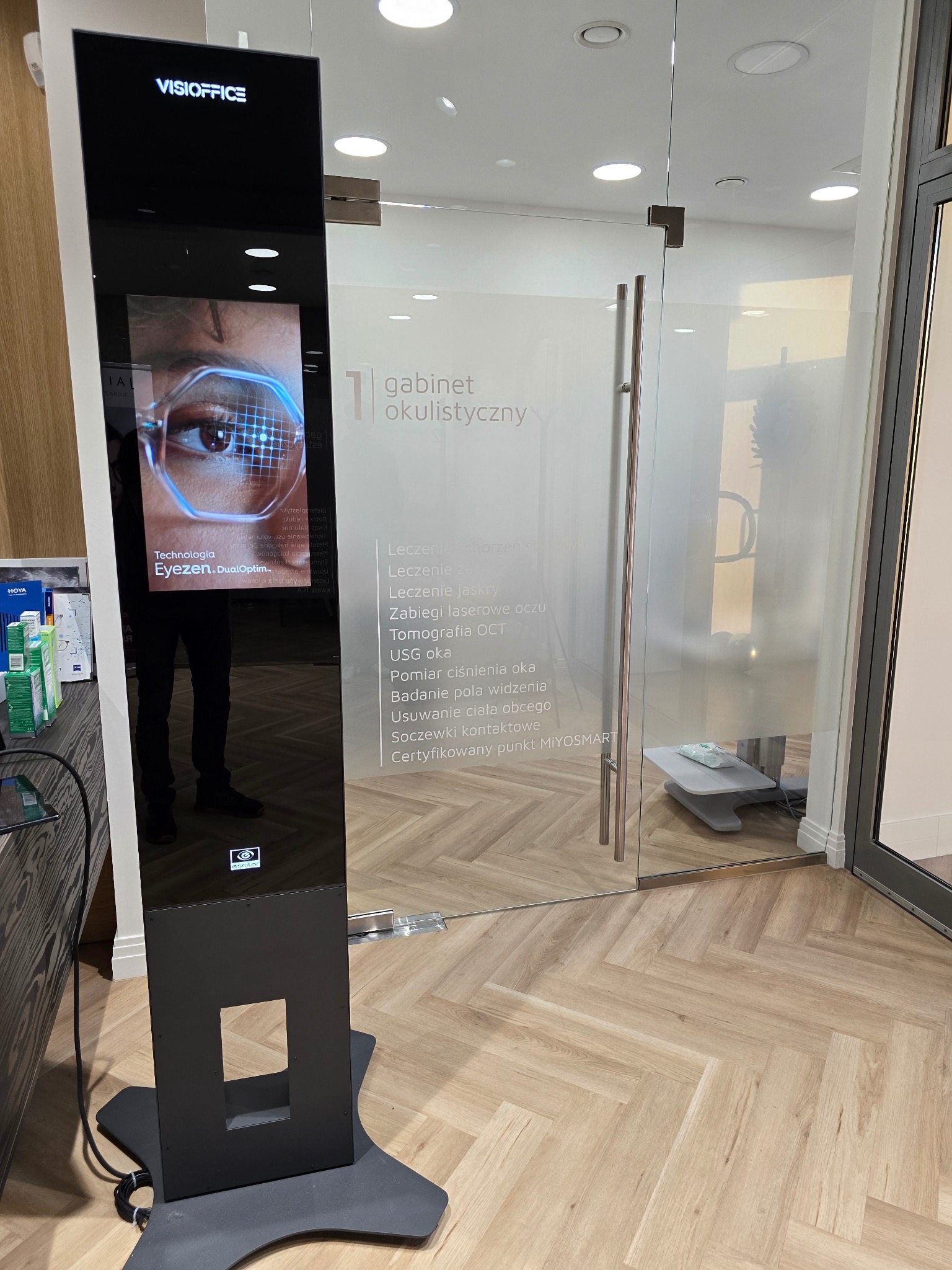 Elegancki gabinet okulistyczny z nowoczesnym ekranem diagnostycznym VisiOffice prezentującym technologię Eyezen DualOptim, widoczne usługi: leczenie jaskry, tomografia OCT, soczewki kontaktowe.