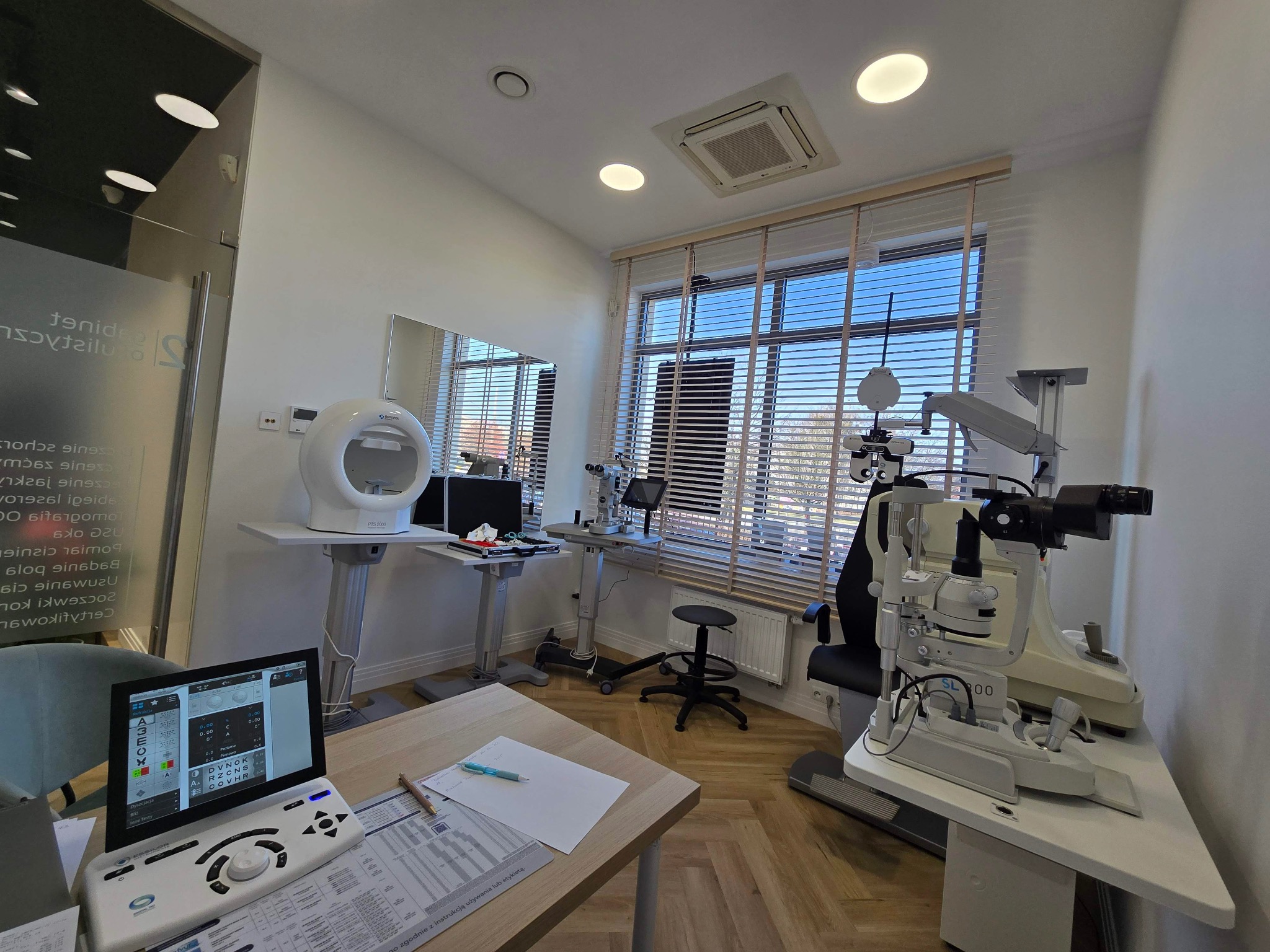 Jasne wnętrze gabinetu lekarskiego z nowoczesnym sprzętem diagnostycznym, w tym tonometrem i lampą szczelinową, oraz komputerem z oprogramowaniem do badania wzroku na pierwszym planie.