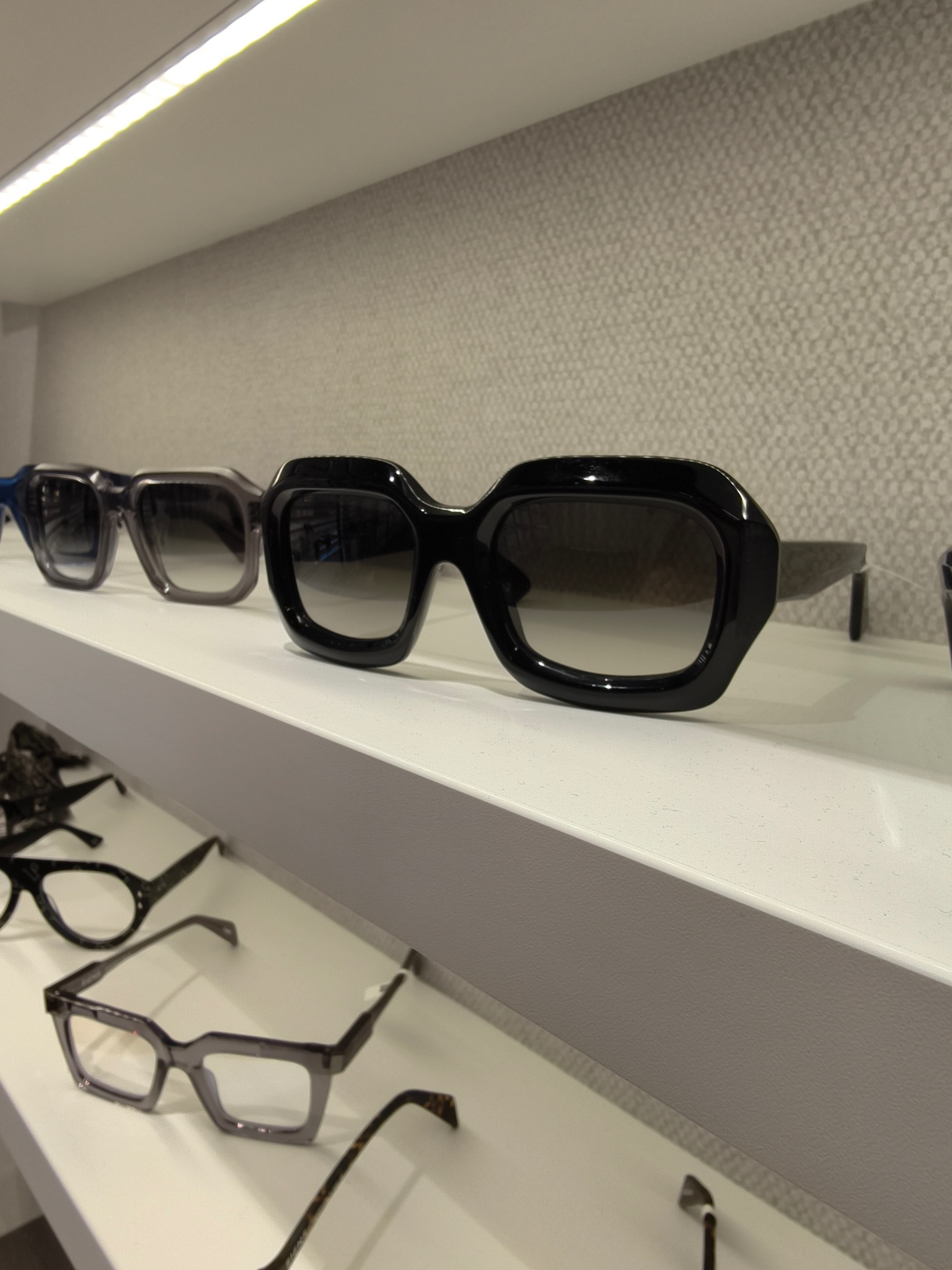 Rząd okularów korekcyjnych i przeciwsłonecznych na białej półce w salonie optycznym, z czarnymi, grubymi oprawkami na pierwszym planie.