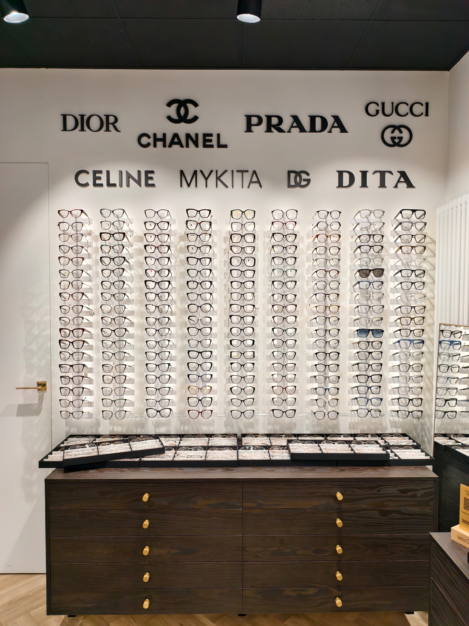 Ekspozycja okularów korekcyjnych i przeciwsłonecznych różnych marek (Dior, Chanel, Prada, Gucci, Celine, Mykita, Dita) na białej ścianie w salonie optycznym, widoczne szuflady z dodatkowymi oprawkami.