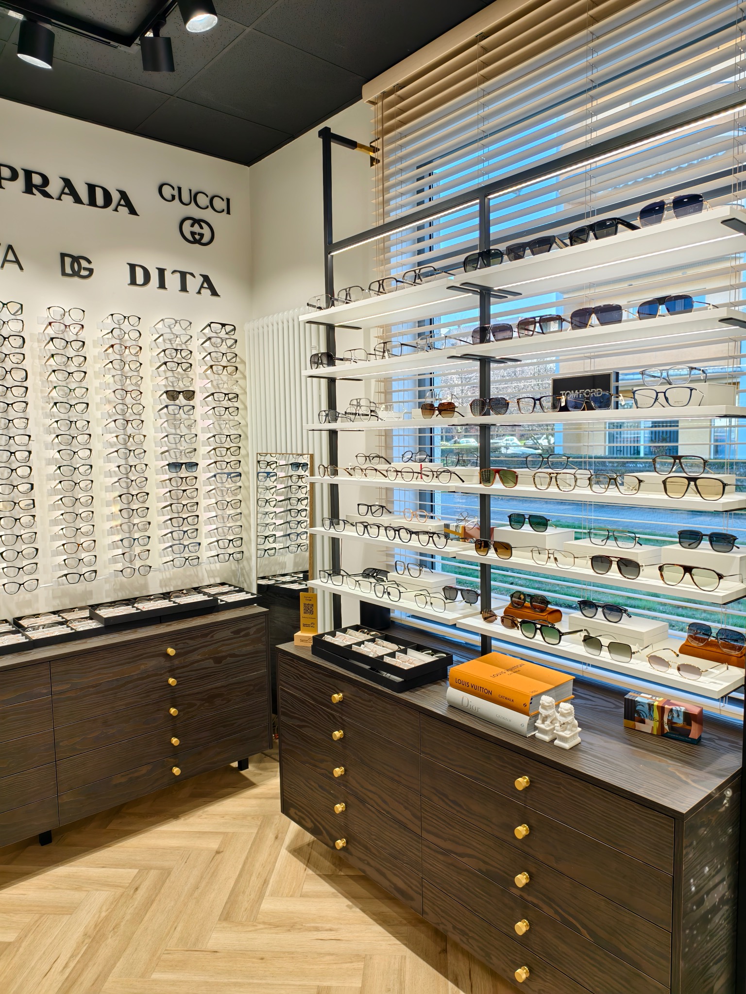 Salon optyczny: półki z okularami korekcyjnymi i przeciwsłonecznymi różnych marek, w tle logo Prada, Gucci, Dita, komody z ciemnego drewna z złotymi gałkami, parkiet w jodełkę.