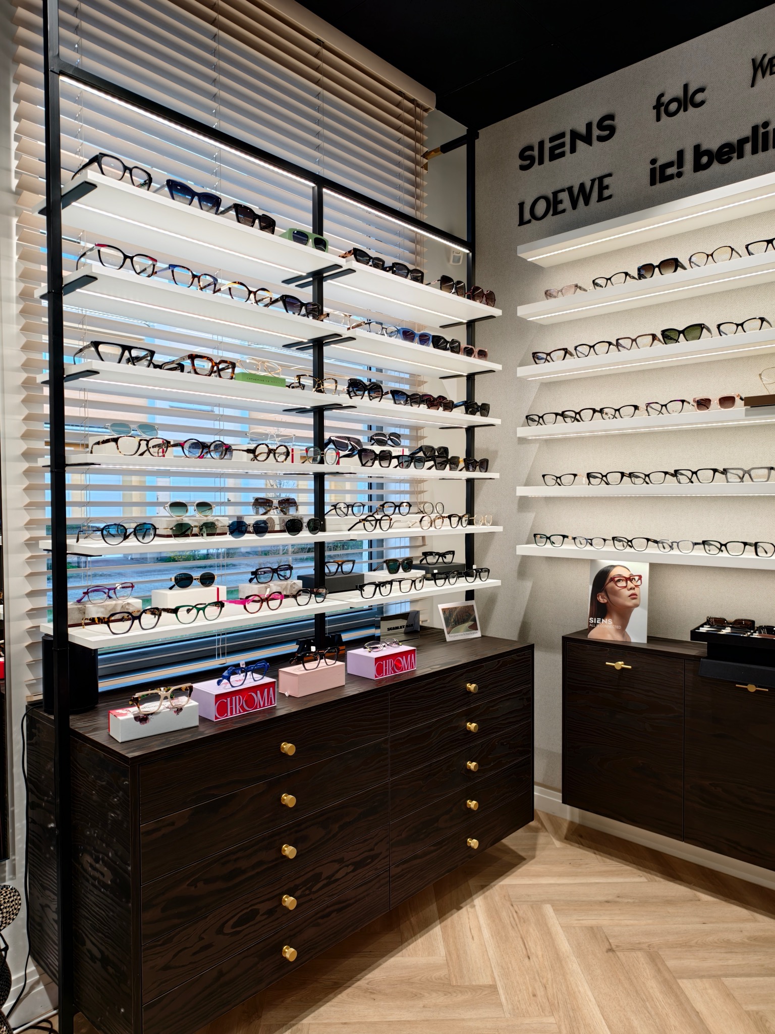 Salon optyczny z ekspozycją okularów na półkach z podświetleniem LED, ciemne drewniane komody z jasnymi gałkami, parkiet ułożony w jodełkę oraz logo marek okularów na ścianie.